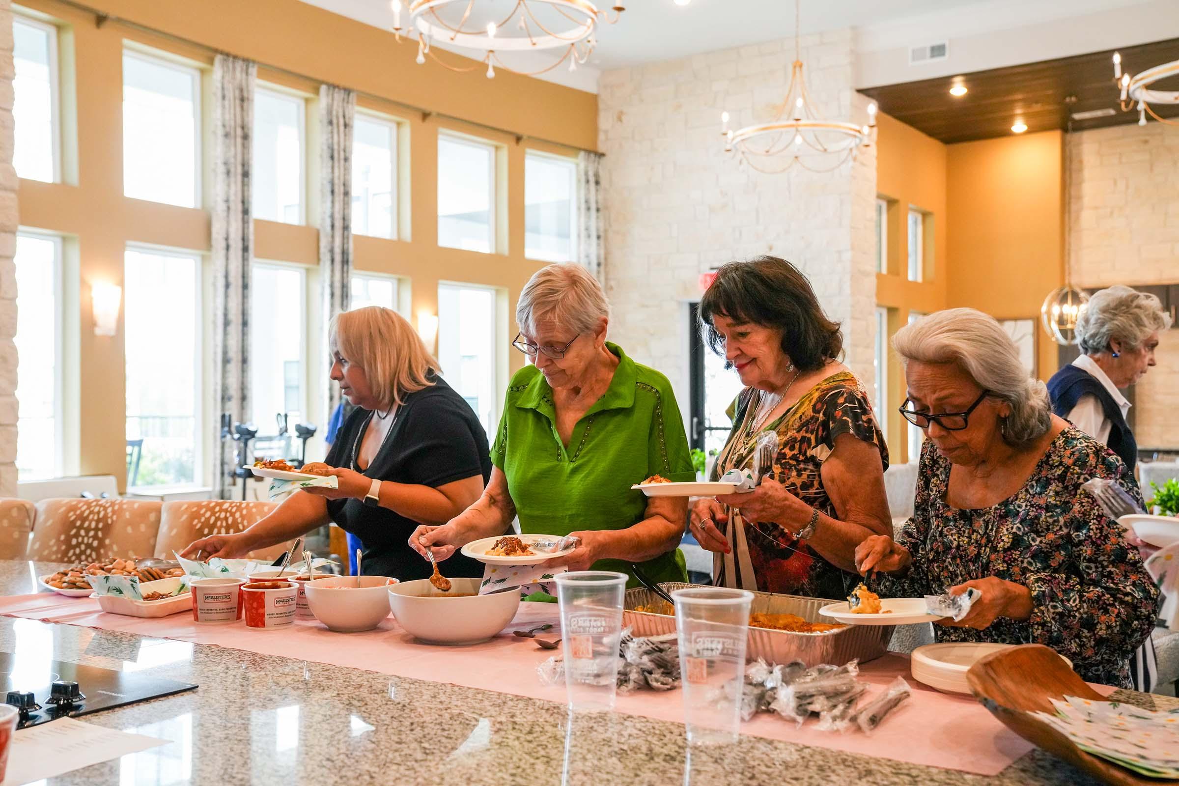 A group of four older women are serving themselves food at a buffet table in a well-lit dining area. Two women are holding plates with food, while the others are preparing to fill their plates. The setting features elegant décor, large windows, and a relaxed atmosphere.