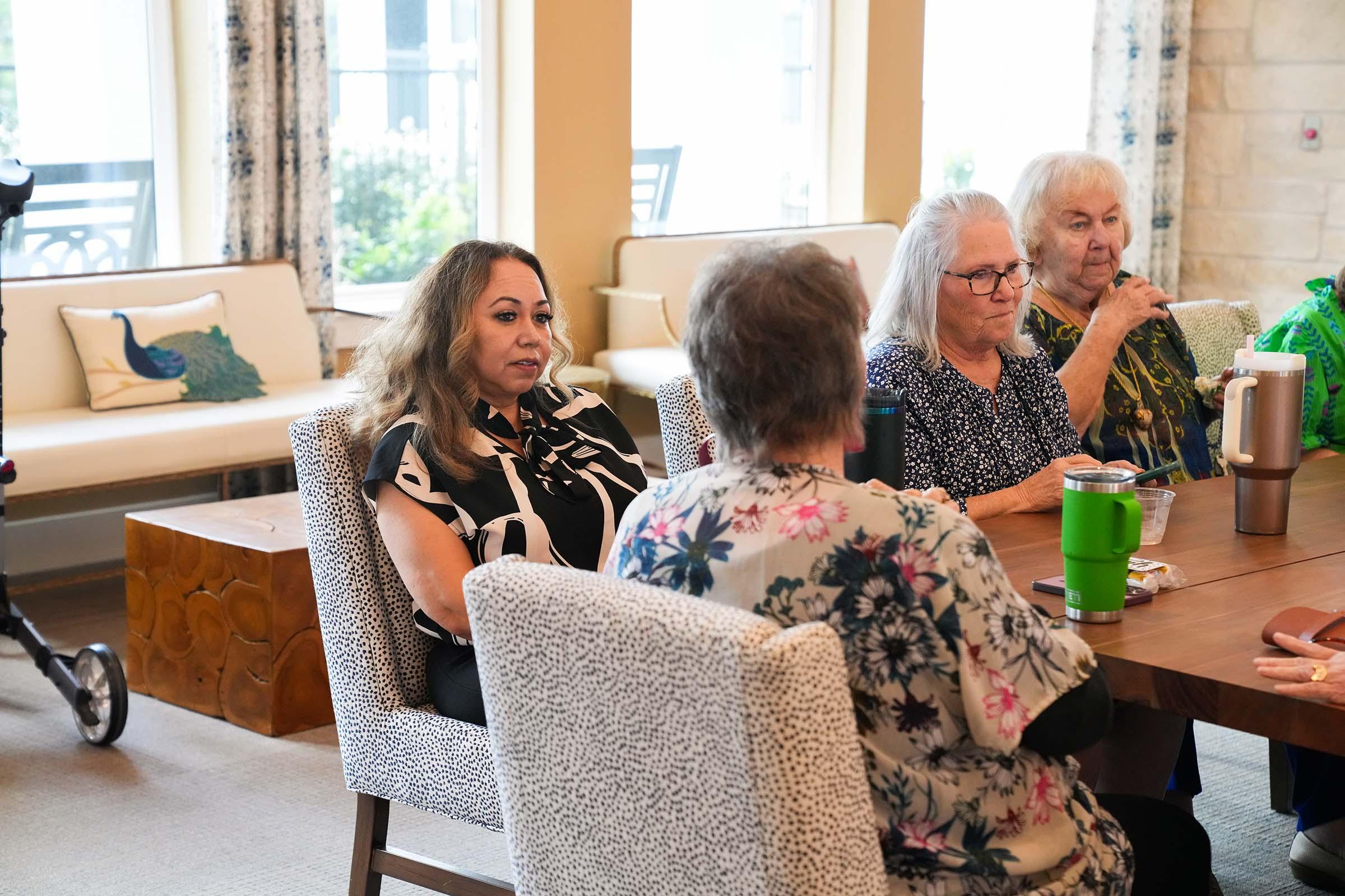 A group of four women sitting around a table in a bright, cozy room. One woman with long hair is engaged in conversation, while the others, including two older women, are listening attentively. The space features large windows and comfortable seating, creating an inviting atmosphere.
