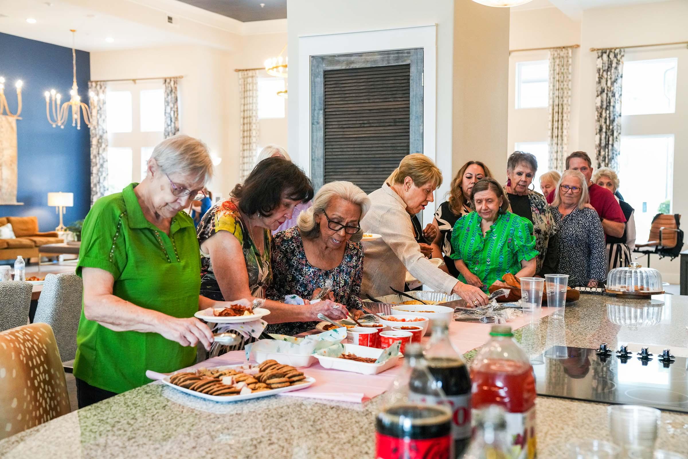 A lively gathering of people at a kitchen countertop, joyfully preparing and serving food. Participants of various ages are engaged in the activity, with an array of dishes and snacks displayed. The setting features modern decor and bright lighting, creating a warm and inviting atmosphere.
