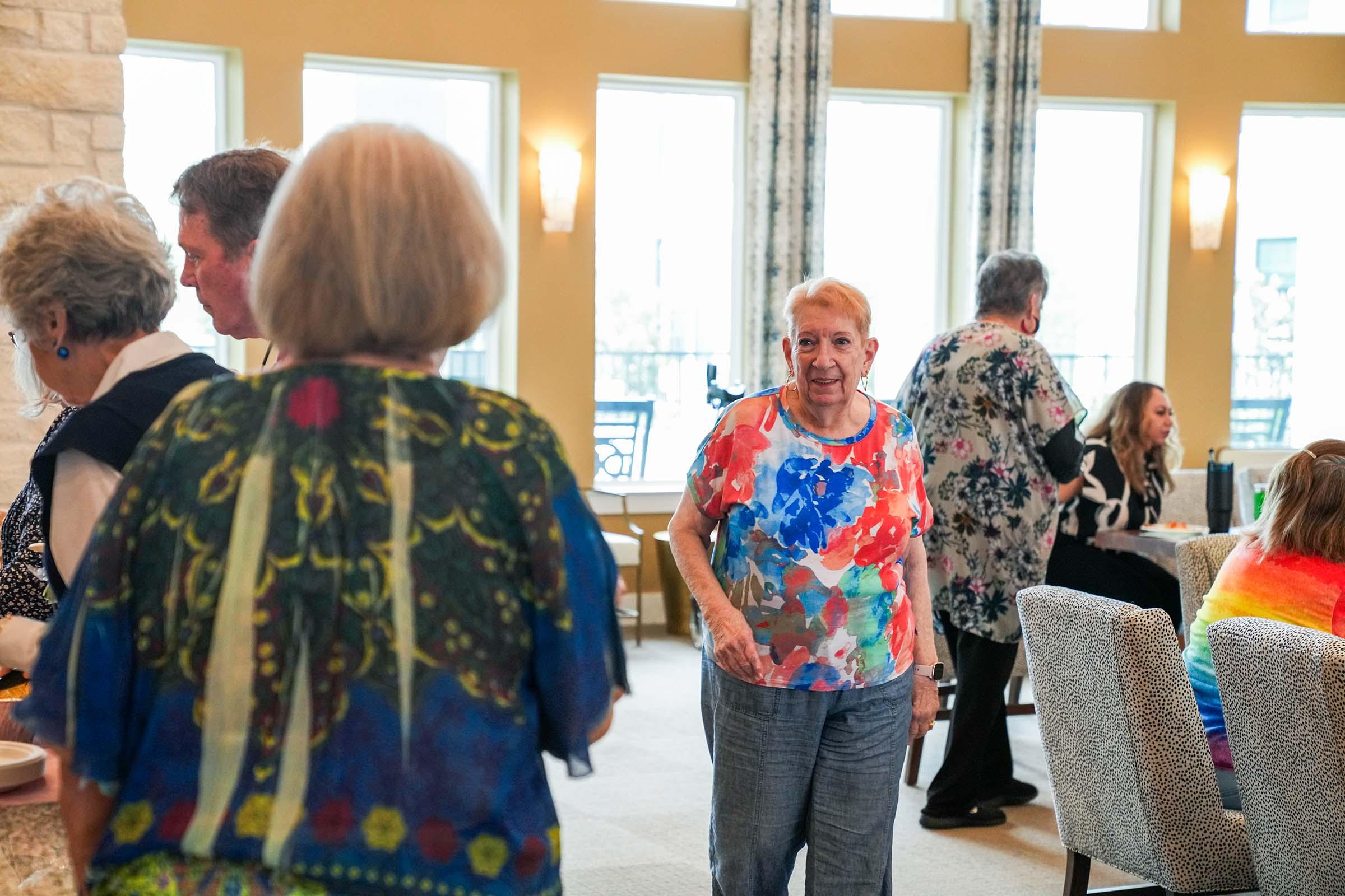 A bright and lively indoor gathering with people of various ages enjoying a social event. An older woman in a colorful floral shirt smiles and interacts with others, while several people in the background share conversation. The setting features large windows and light decor, creating a warm atmosphere.