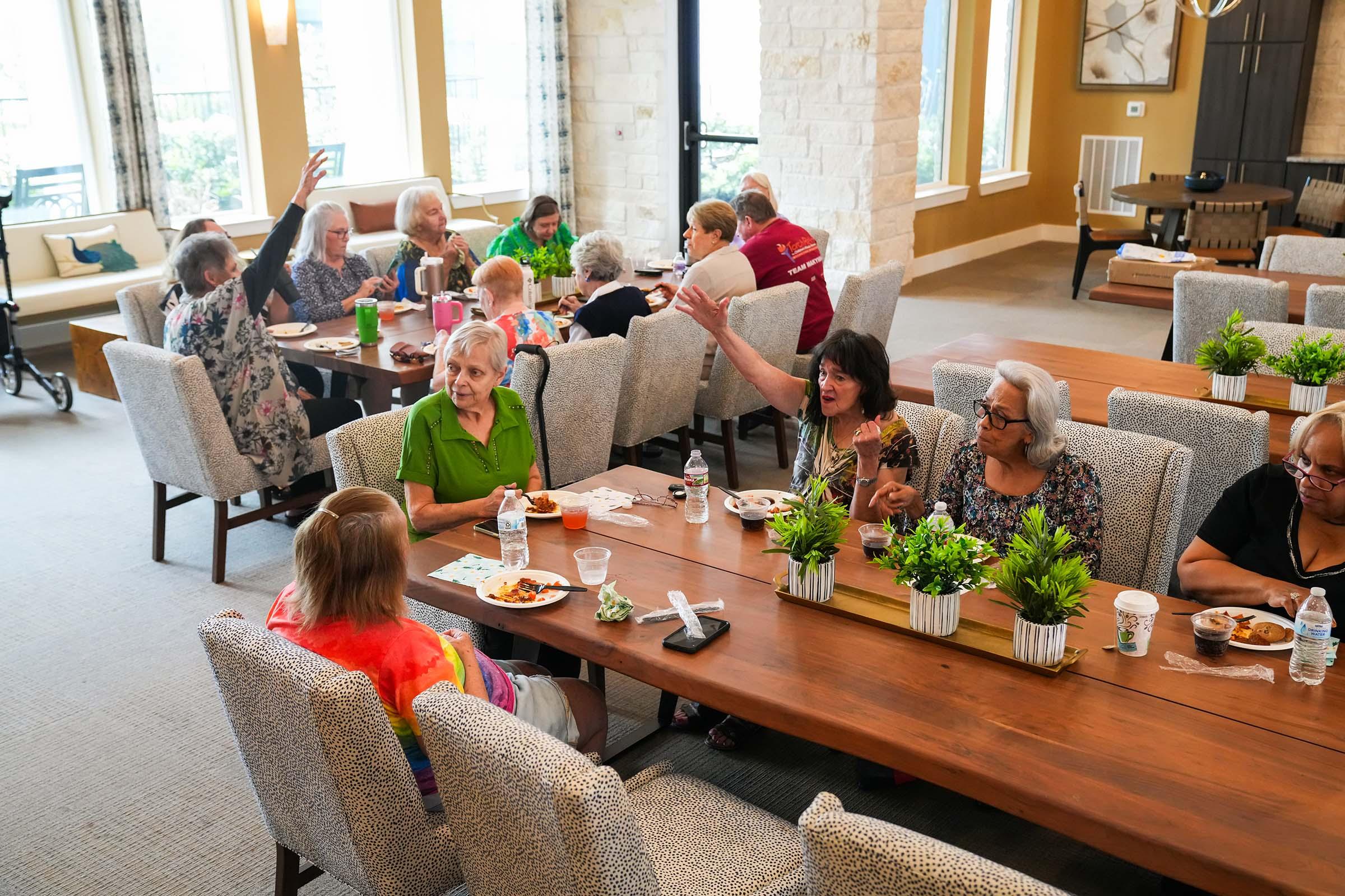 A lively group of seniors enjoying a meal together in a spacious dining area. Several people are engaged in conversation, with some gesturing animatedly. The table is set with food and drinks, and greenery adds a touch of warmth to the setting. The atmosphere appears cheerful and communal.