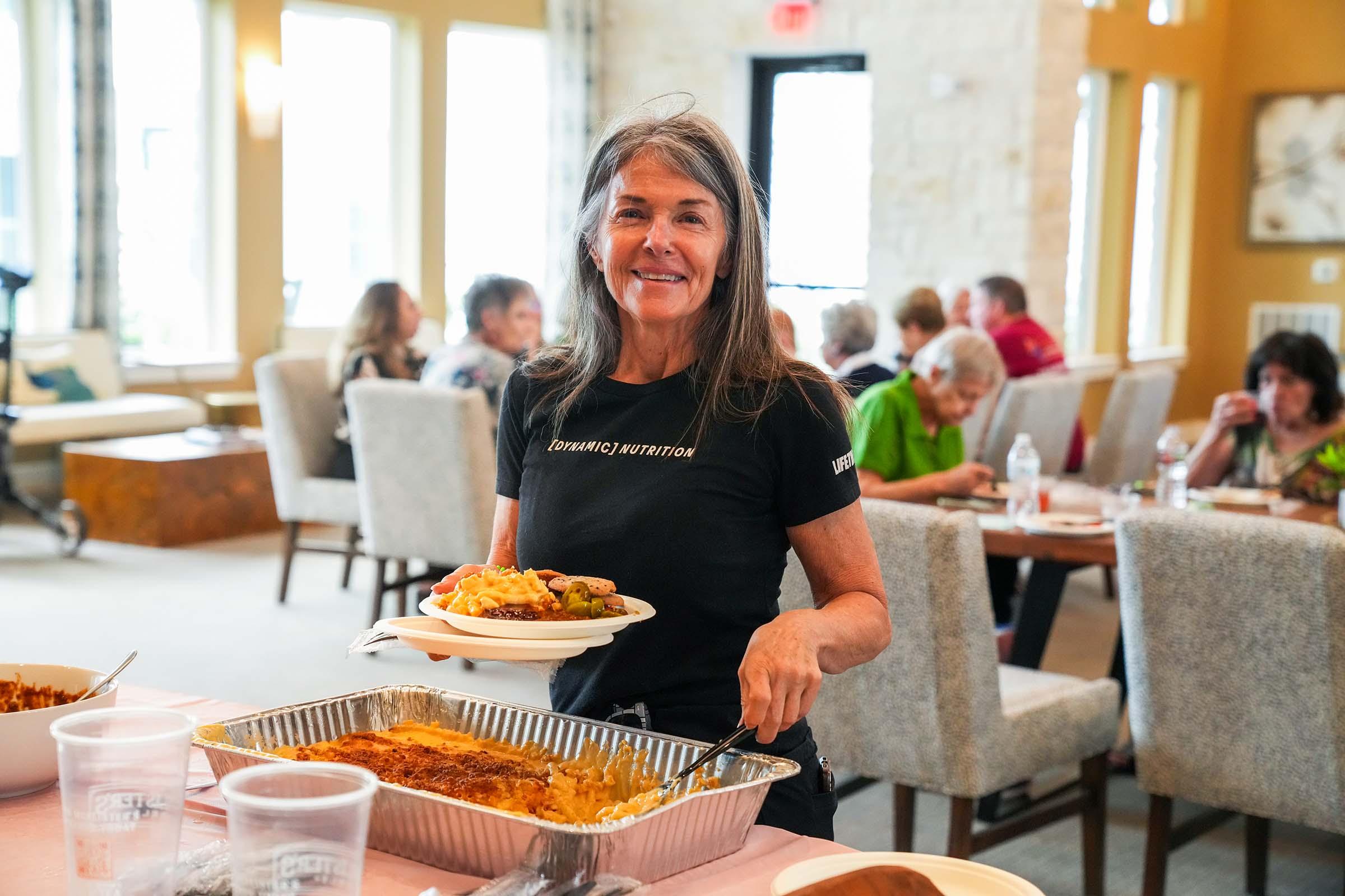 A smiling woman holds a plate of food in a community dining space, dressed in a black t-shirt. In the background, others are seated at tables enjoying their meals. The setting has large windows and light-colored decor, creating a welcoming atmosphere.