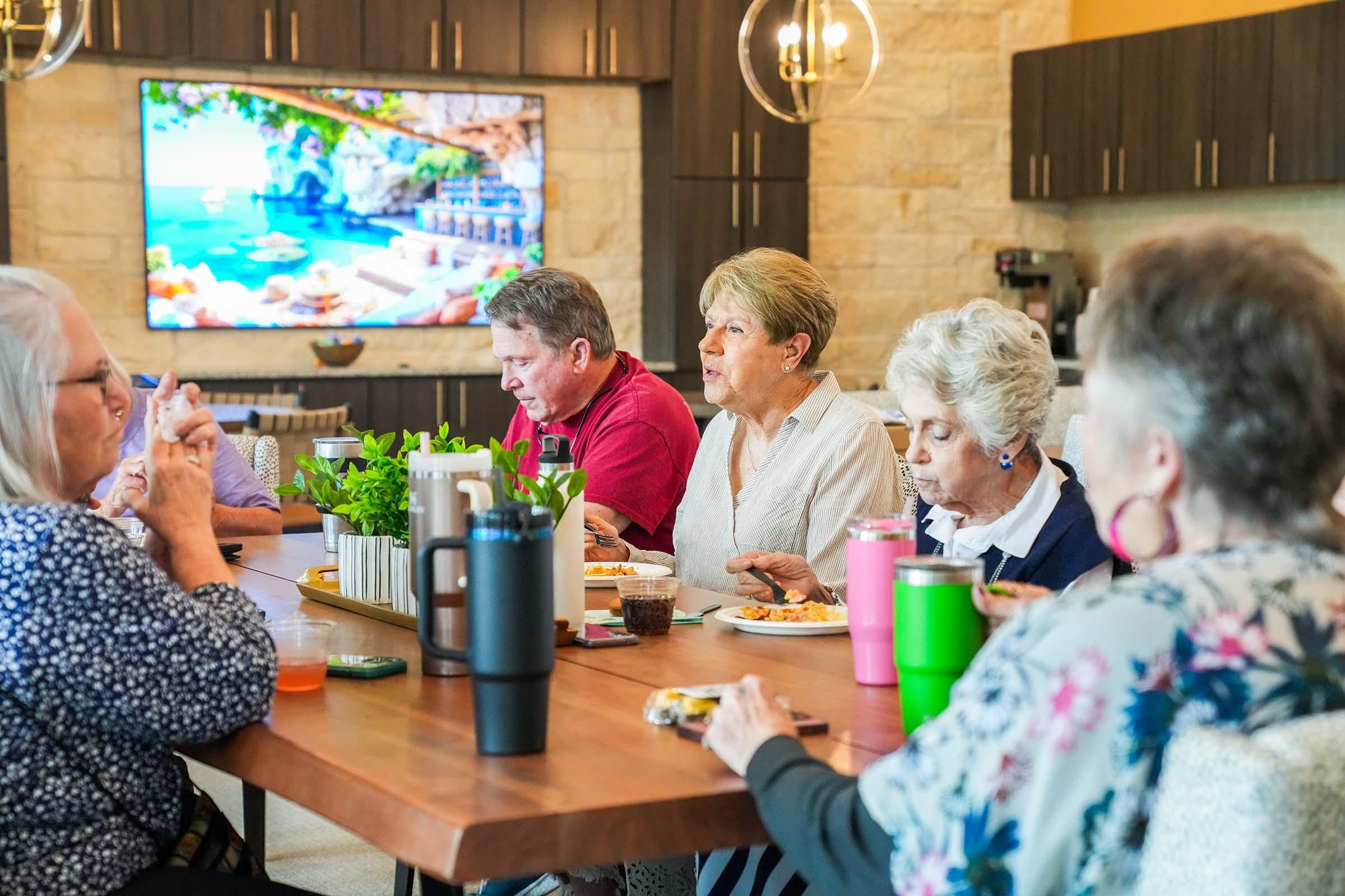 A group of six people enjoying a meal around a wooden table in a modern kitchen setting. There is a colorful screensaver of a tropical beach displayed on a large TV in the background. The atmosphere appears casual and cheerful, with plates of food and drinks present on the table.