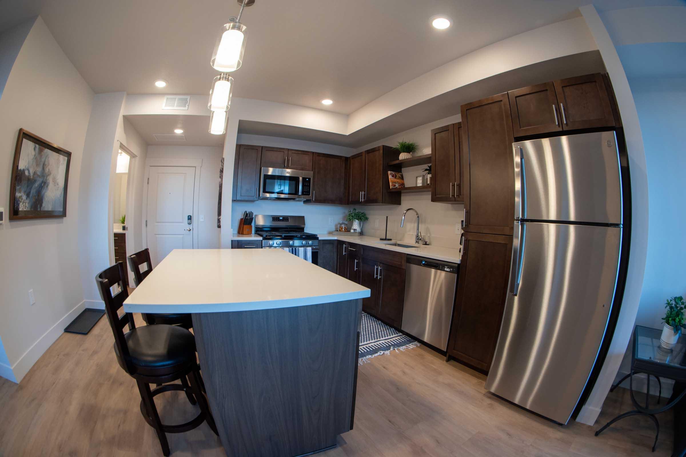 Modern kitchen with wooden cabinets, stainless steel appliances including a refrigerator and microwave, and a large island with seating for two. Soft overhead lighting illuminates the space, which features light-colored walls and wood flooring. A decorative rug adds texture to the area.