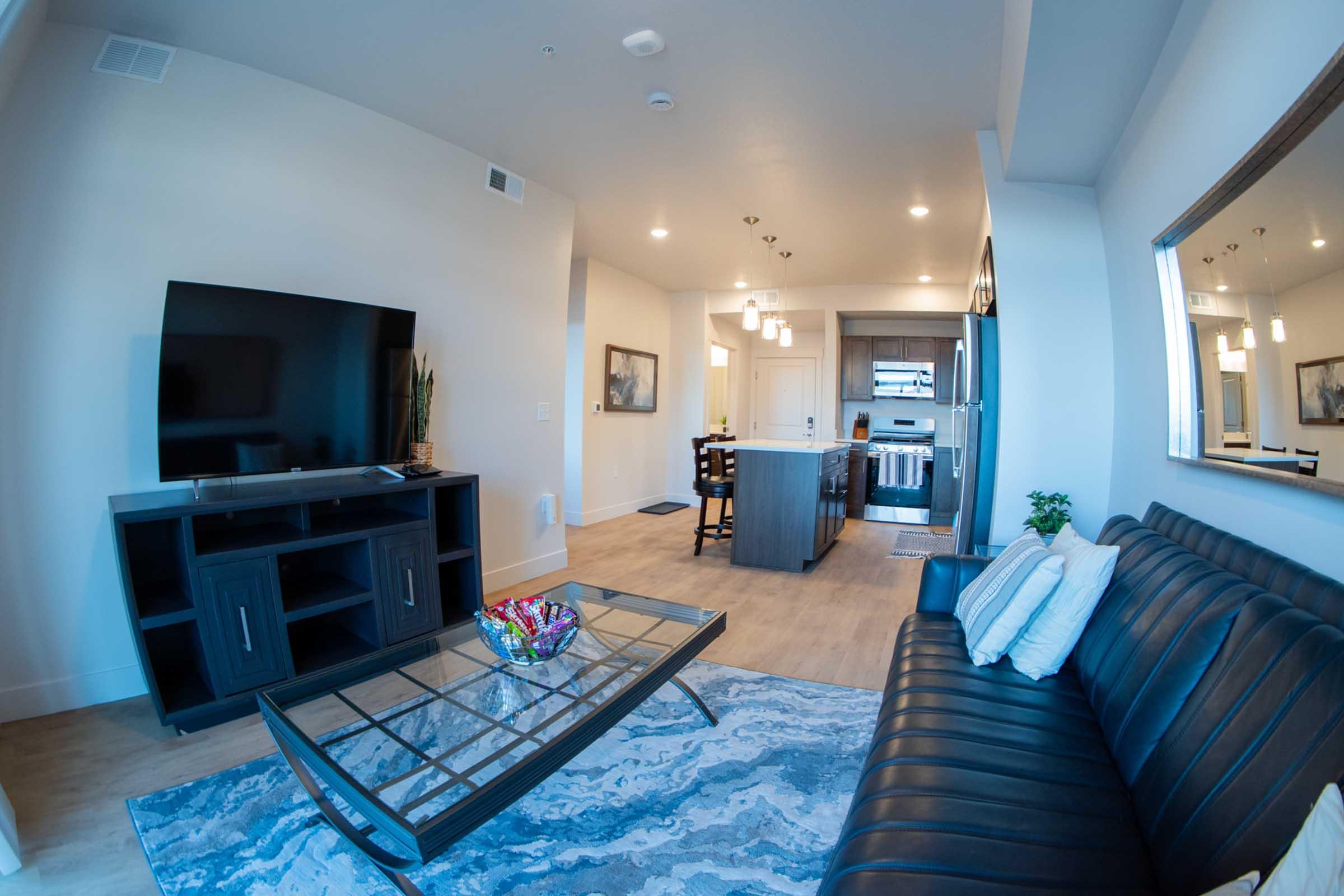 A modern living room featuring a black leather sofa, a glass coffee table with a colorful bowl, and a large flat-screen TV on a dark wooden stand. The background shows a dining area with bar stools and a kitchen. Natural light illuminates the space, highlighting the contemporary decor and light wood flooring.