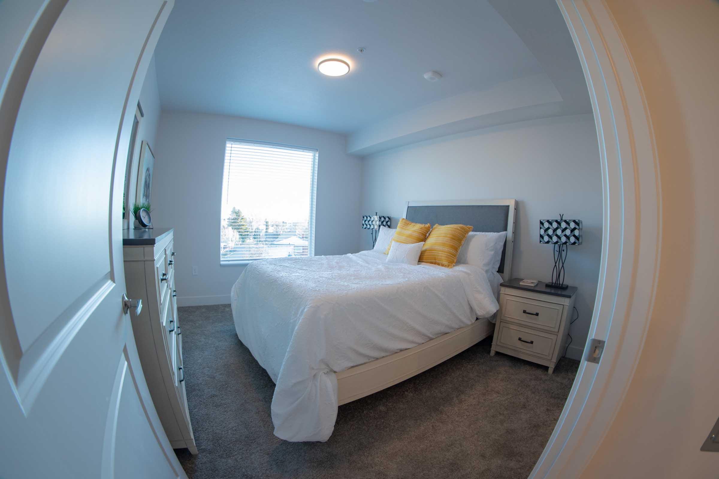 A well-lit bedroom featuring a cozy queen-sized bed with white bedding and yellow decorative pillows. There are two bedside lamps on small tables, a window with natural light, and a view of the outdoors. The walls are painted in a light color, and plush carpeting adds warmth to the space.