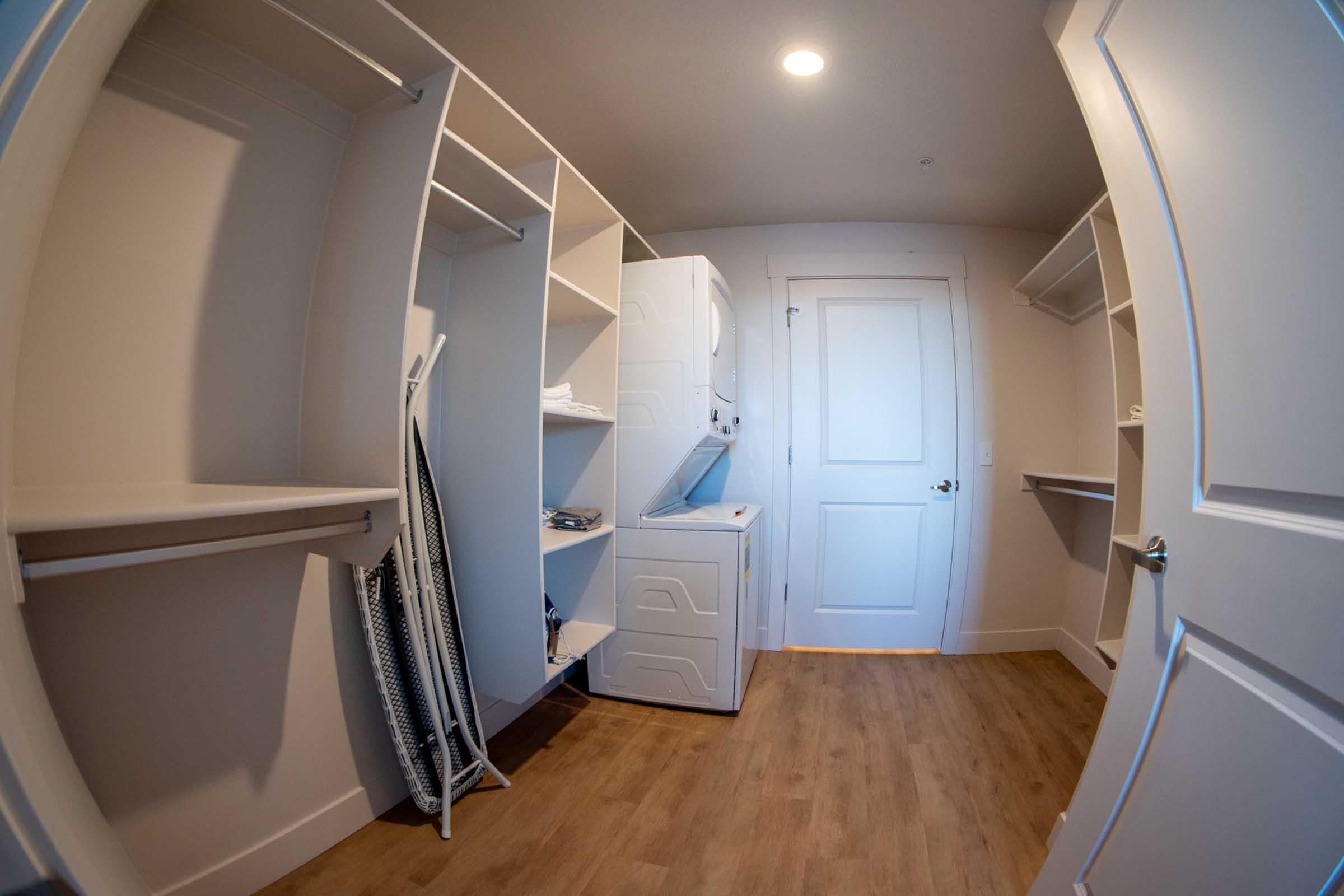 A well-organized laundry room featuring white shelving units on both sides, a stacked washer and dryer against one wall, and a door leading to another room. The floor is wooden, and the walls are painted in a light color, creating a spacious and bright environment.