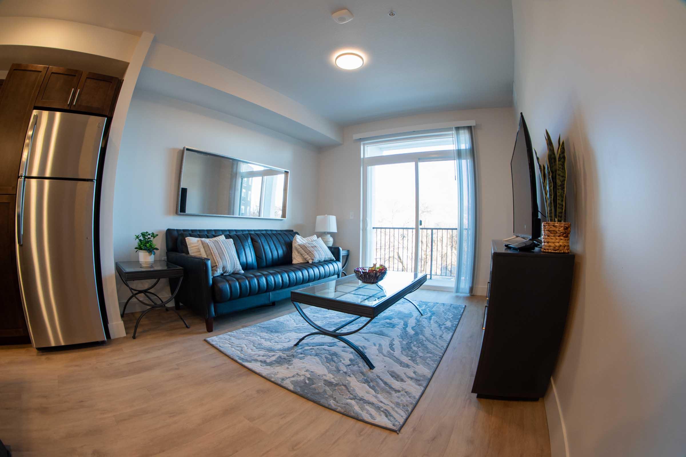 A modern living room featuring a dark blue leather couch, a glass coffee table on a patterned rug, and a wall-mounted TV. A large window allows natural light to fill the space, with a view of an outdoor area. The room includes a stainless steel refrigerator and decorative plants on a side table.