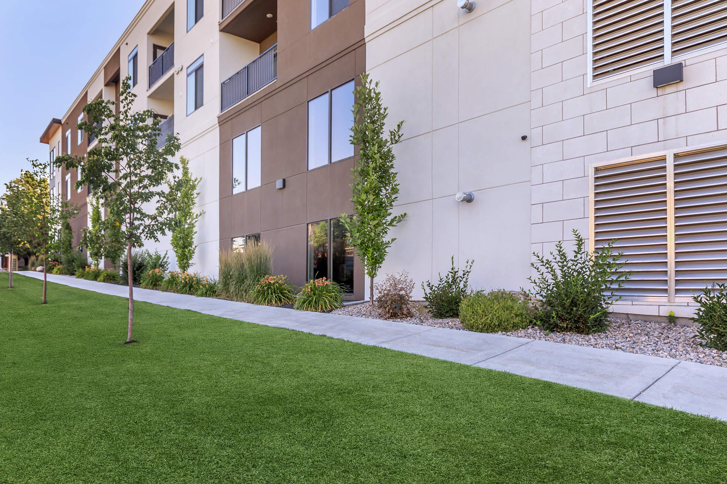 A well-maintained outdoor area featuring a modern apartment building with a manicured lawn. The scene includes neatly trimmed shrubs and small trees lining a paved walkway, providing a clean and inviting ambiance. Sunlight bathes the area, enhancing the vibrant greenery and architectural details of the building.