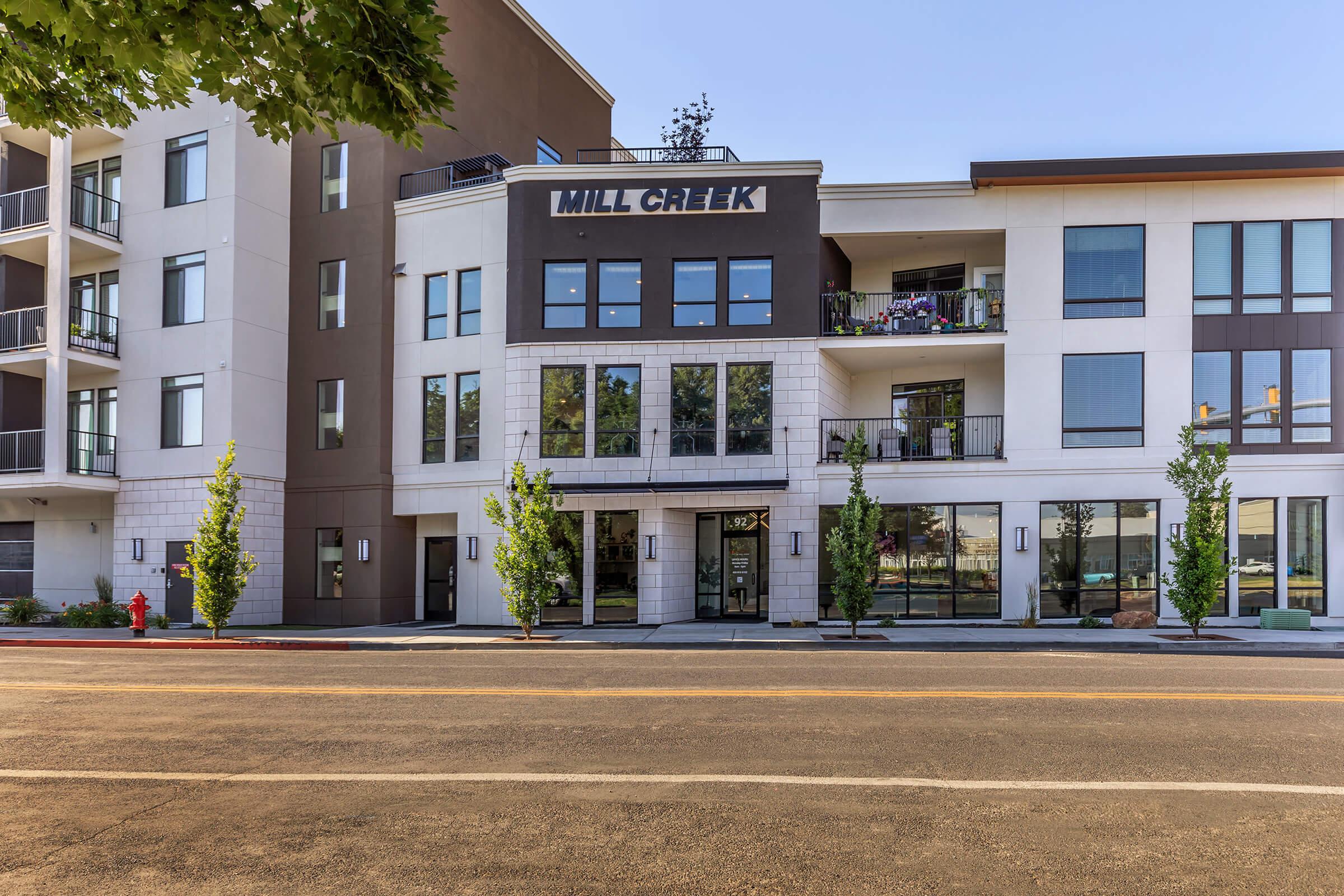 A modern multi-story building with the name "MILL CREEK" prominently displayed on the front. The architecture features a mix of textures and materials, large windows, and balconies with greenery. In front, there are small trees lining the sidewalk, and a fire hydrant is visible. The scene is bathed in natural light.