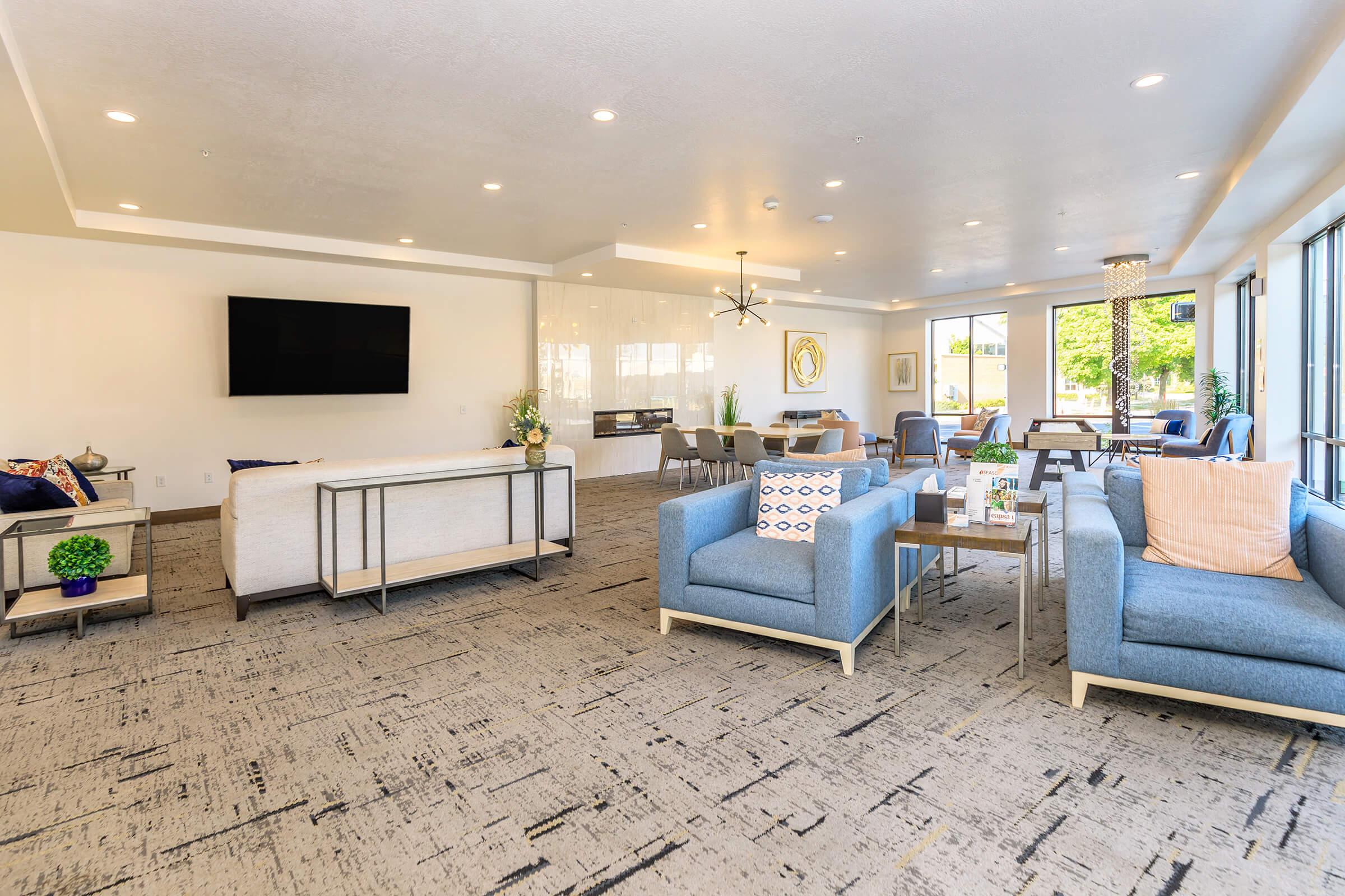 A modern, inviting lounge area featuring light blue armchairs, a white sofa, and stylish decor. Large windows allow natural light, and there's a flat-screen TV mounted on the wall. Decorative tables and fresh flowers accentuate the space, creating a cozy atmosphere for relaxation or socializing.