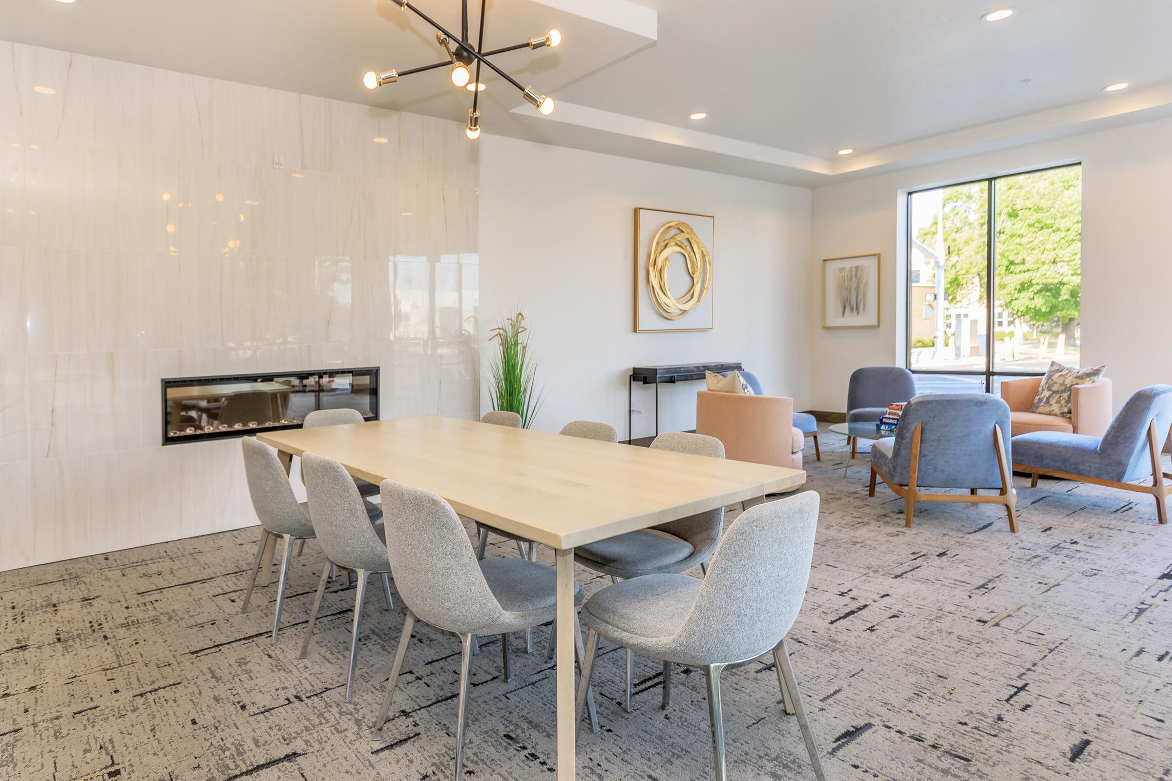 A modern interior space featuring a long wooden dining table surrounded by gray chairs. In the background, there is a stylish wall-mounted fireplace and large windows letting in natural light. The decor includes abstract art and potted plants, with additional seating in the form of plush chairs.