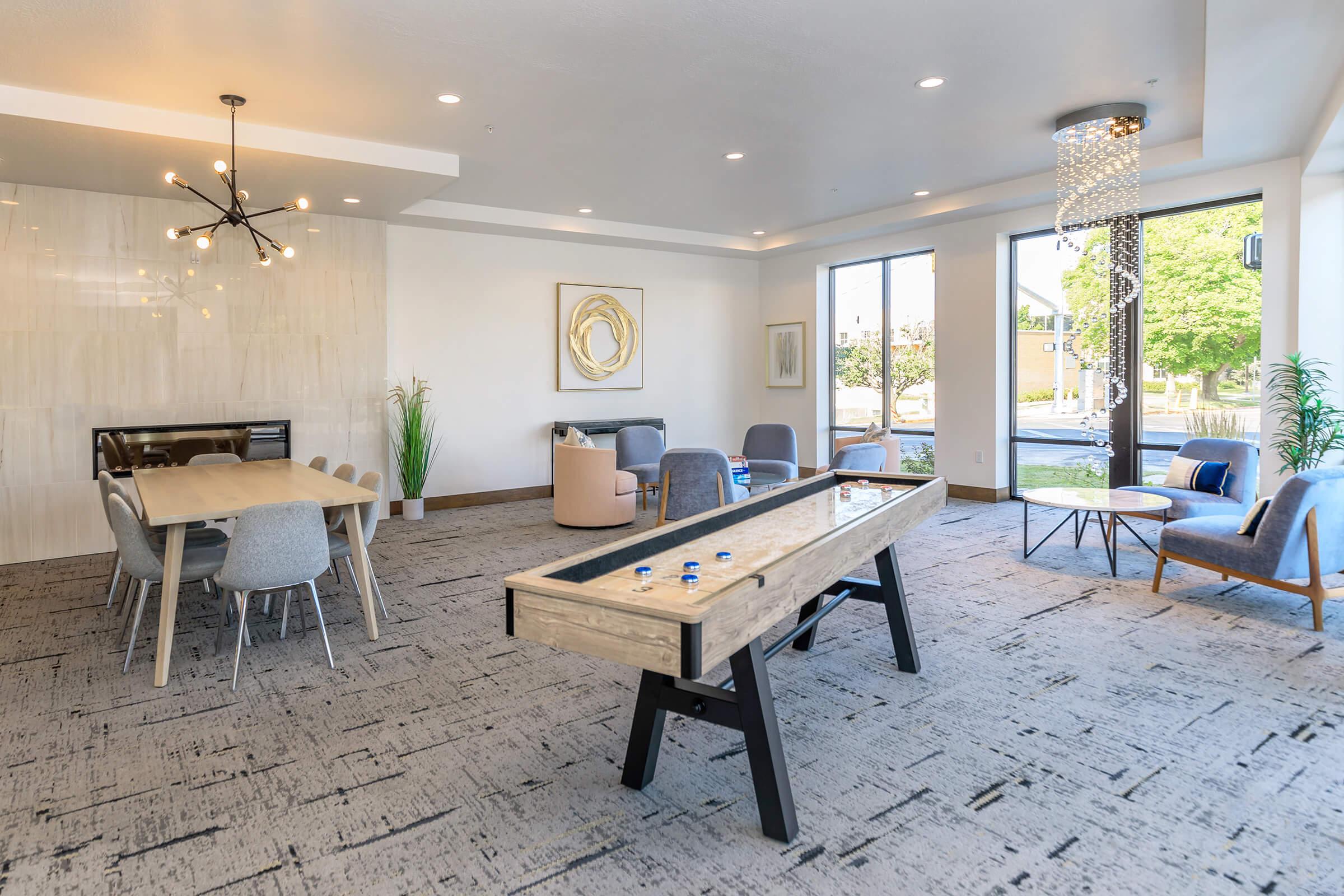 A modern indoor lounge area featuring a shuffleboard table in the center, surrounded by comfortable seating including armchairs and a dining table. Large windows allow natural light to fill the space, highlighting the stylish decor and contemporary lighting fixtures. Green plants add a touch of nature.