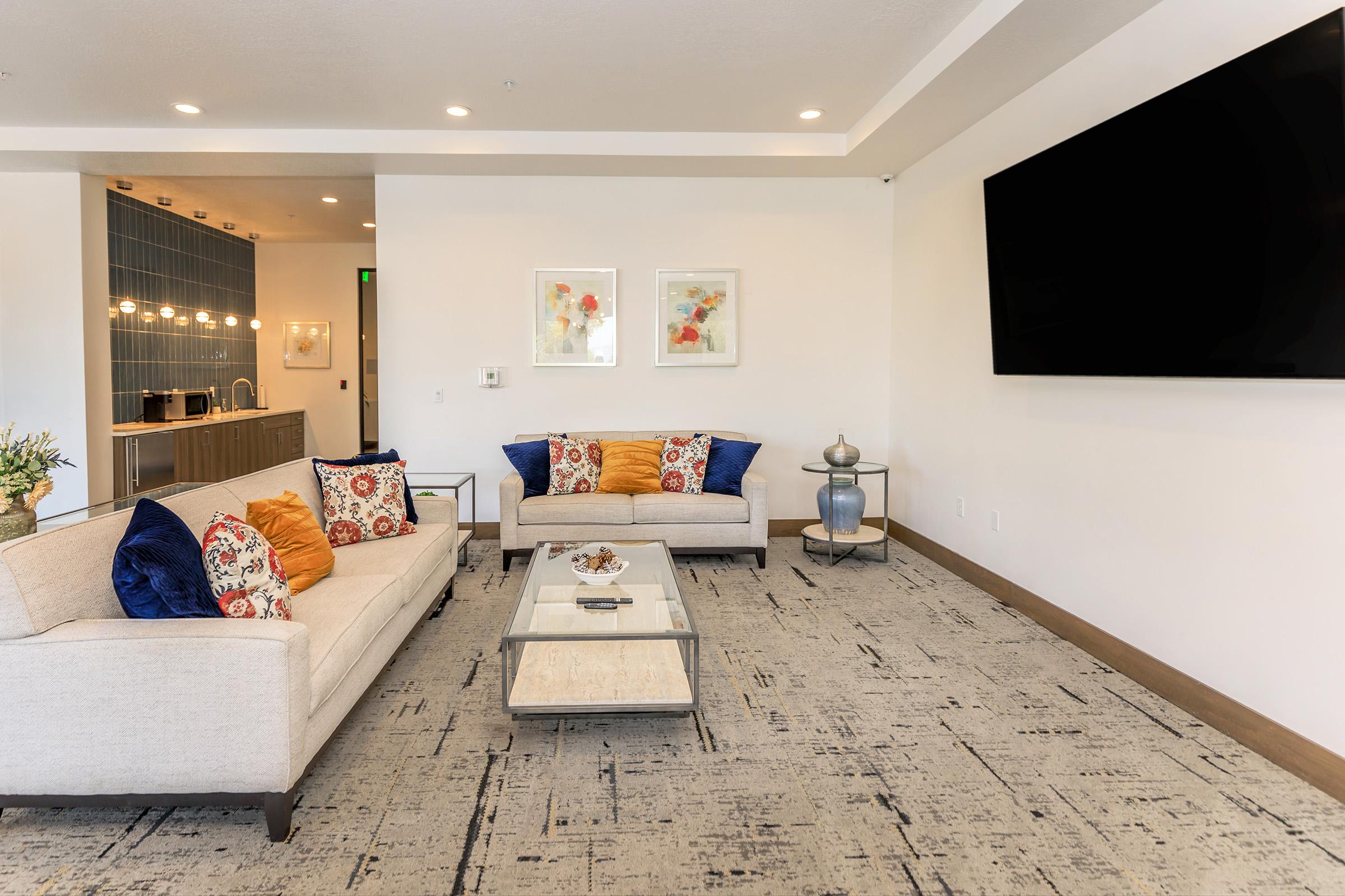 A modern living room featuring two light-colored sofas with decorative pillows, a coffee table in the center, and a large wall-mounted television. The decor includes artwork on the walls, a carpet with an abstract pattern, and a small side table with decorative items. The overall ambiance is inviting and stylish.