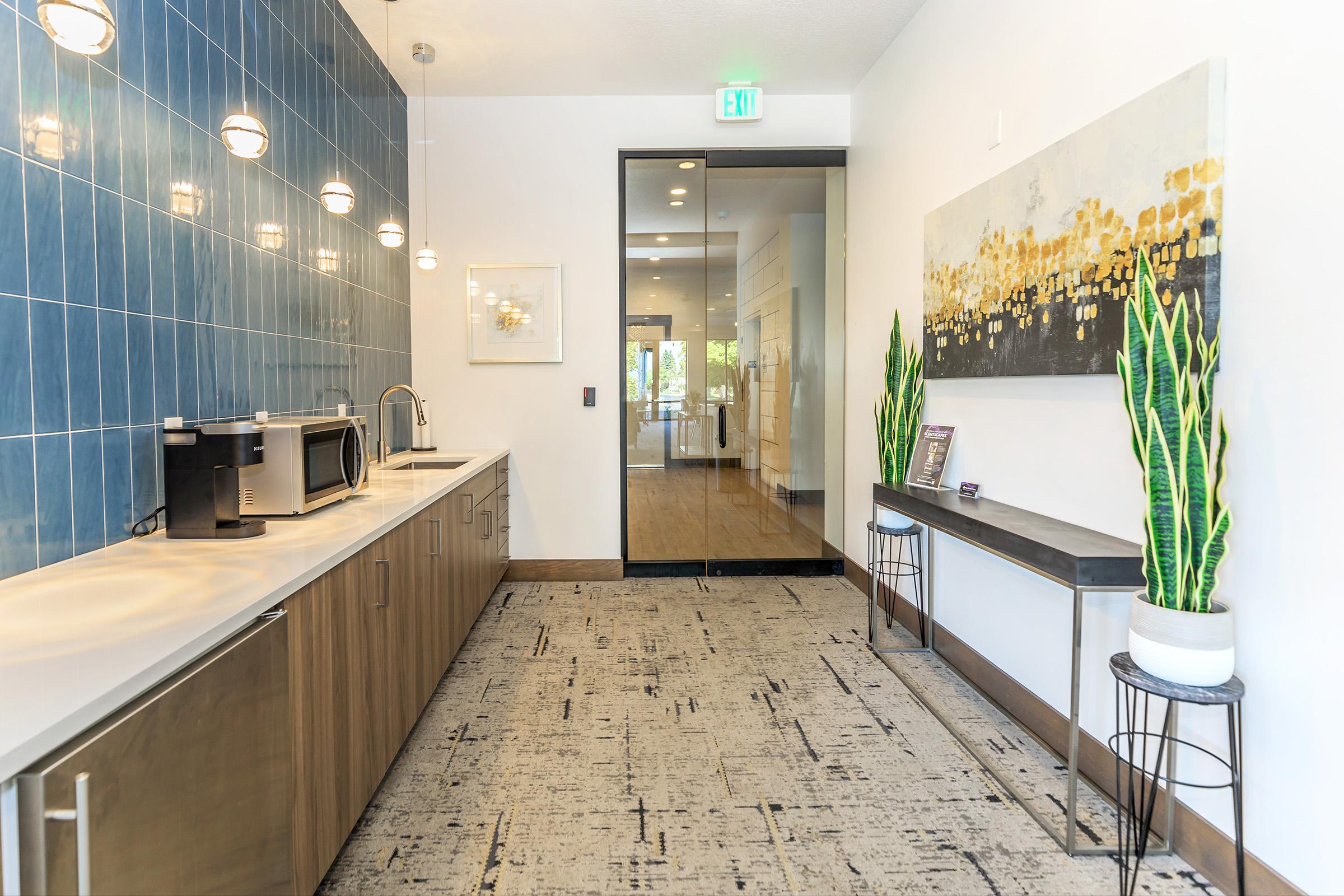 A modern kitchen area featuring a sleek countertop with a microwave, stylish pendant lights, and a large glass door leading to another room. Decorative plants are placed on a console table against a wall adorned with abstract artwork in gold and blue hues. The flooring is textured and contemporary.