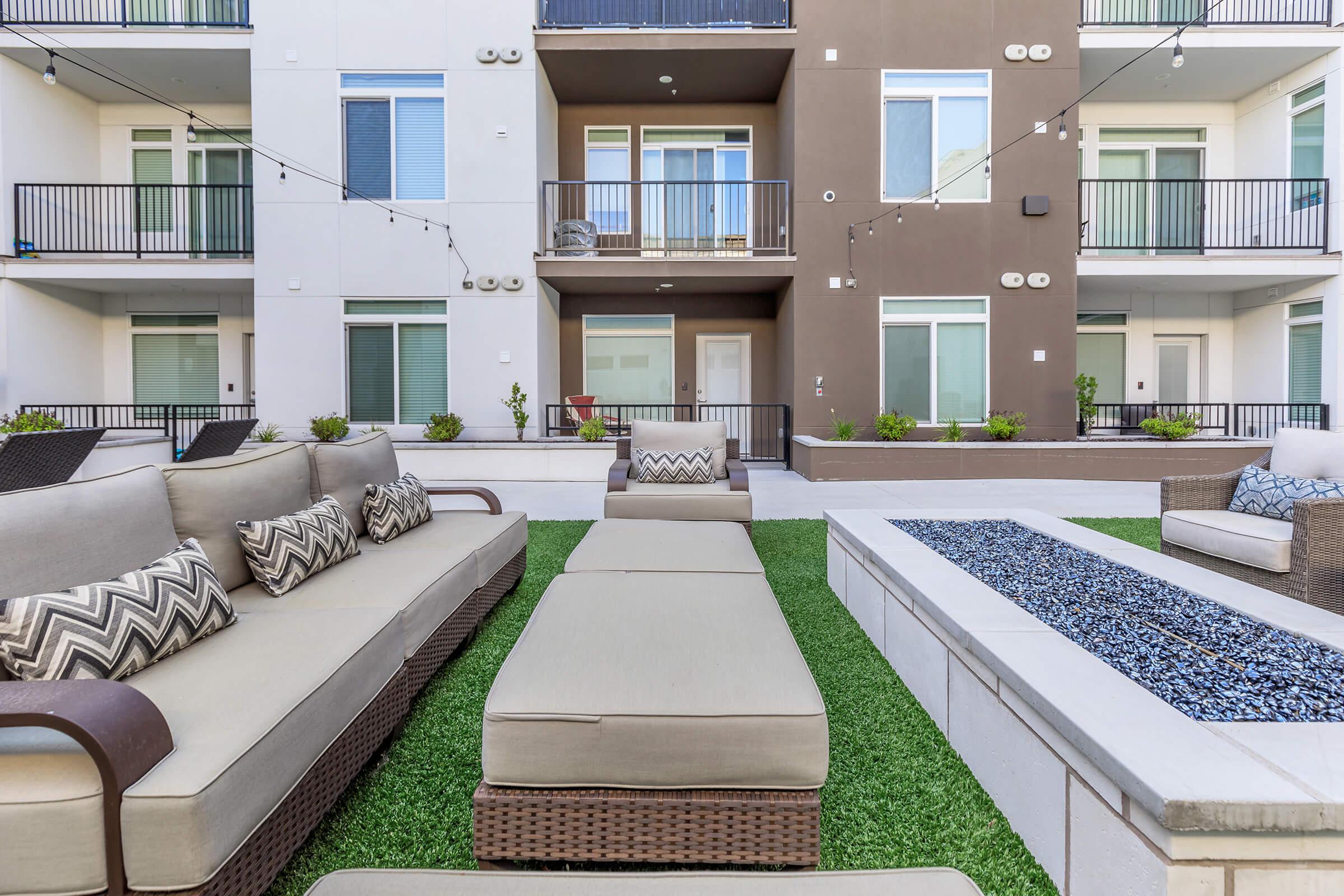 Modern outdoor seating area featuring large, cushioned sofas arranged on green synthetic grass. A stone fire pit with decorative rocks is in the foreground, and apartment building balconies with potted plants are visible in the background. Soft lighting strings hang above for ambiance.