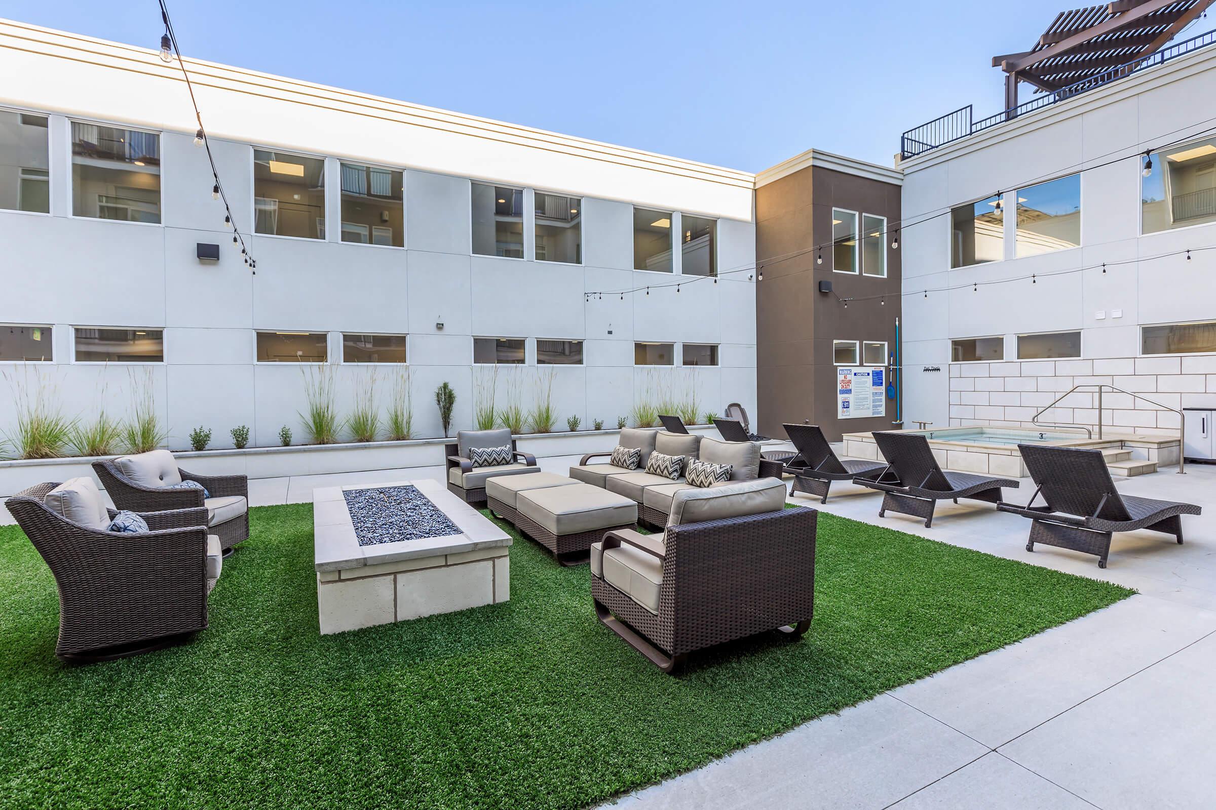 A modern outdoor lounge area featuring comfortable seating on artificial grass, a fire pit in the center, and lounge chairs arranged around the space. The backdrop includes a light-colored building with large windows, creating a welcoming atmosphere for relaxation.