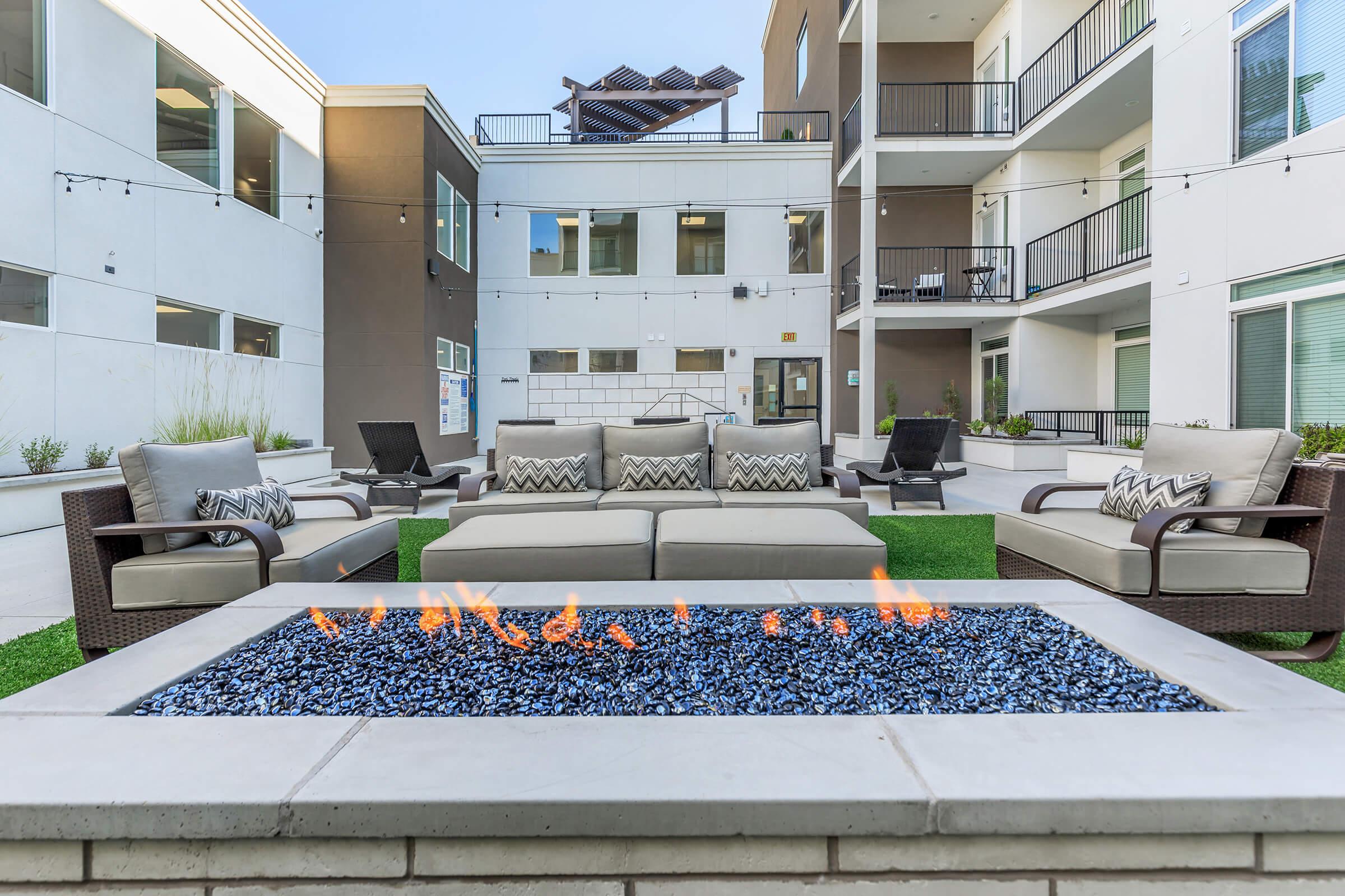 A cozy outdoor lounge area featuring a modern fire pit with blue stones, surrounded by stylish seating on green grass. The backdrop includes a multi-story building with balconies, large windows, and string lights for a warm ambiance. Ideal for relaxation and social gatherings.