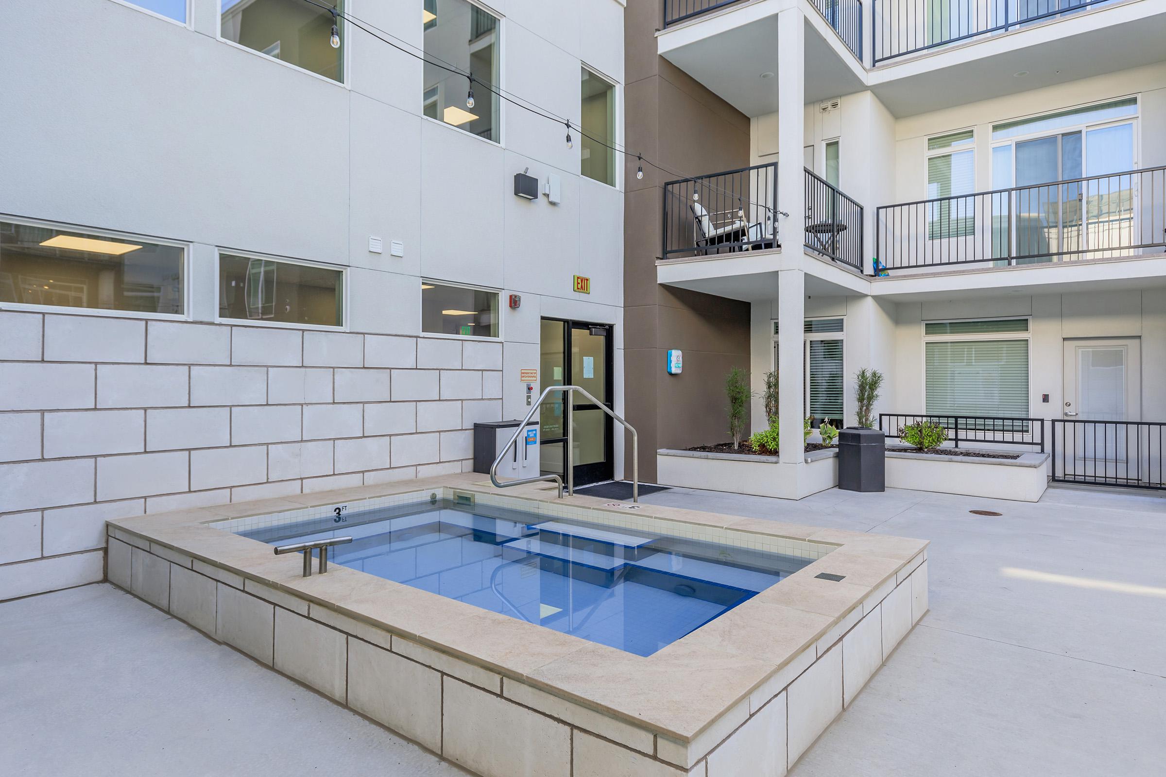 A small, modern outdoor hot tub is situated in an apartment courtyard, surrounded by beige and gray walls. There are several balconies visible with sliding glass doors, and potted plants add greenery. The area is well-lit and clean, suggesting a welcoming space for relaxation.