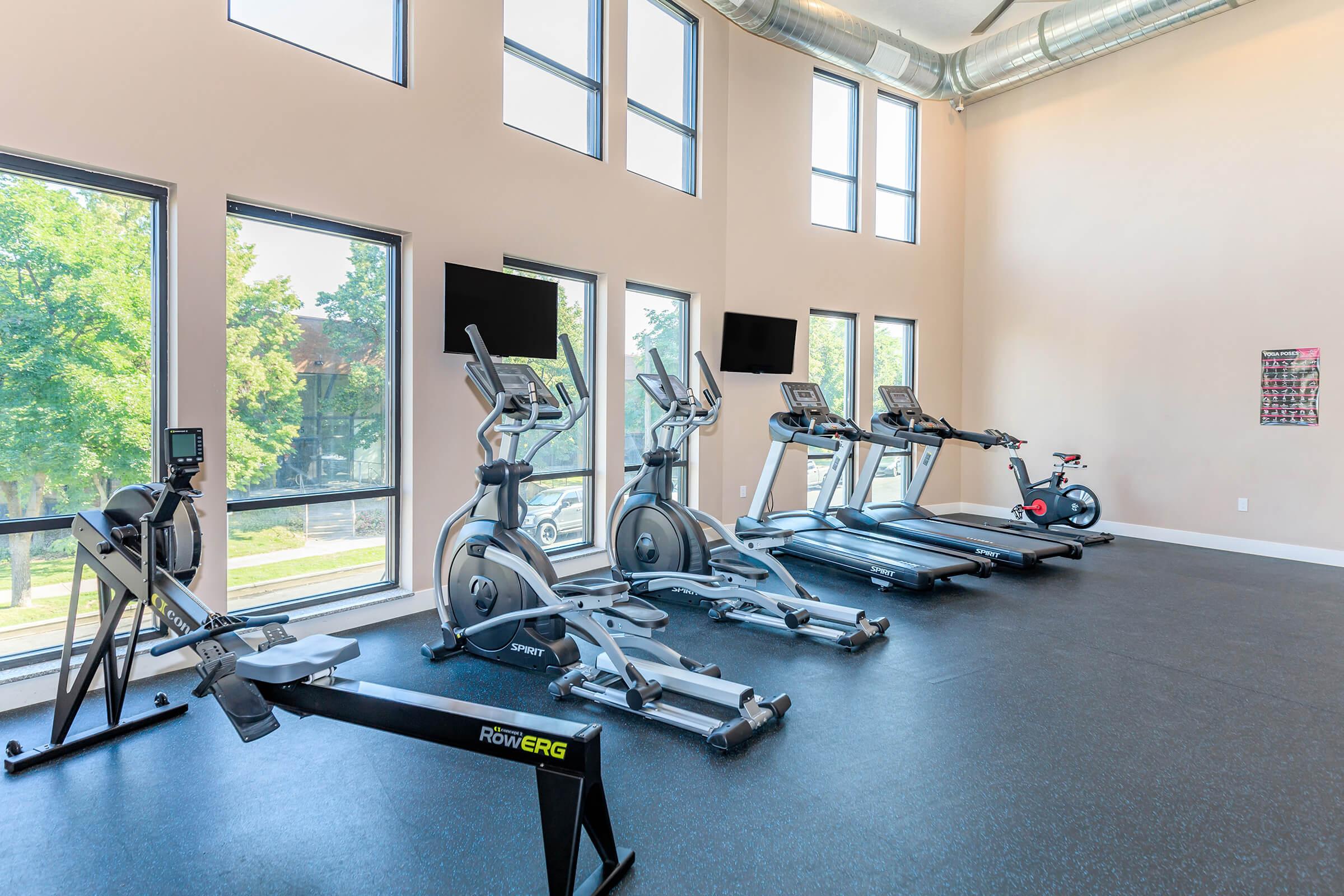 A bright, spacious gym with large windows allowing natural light. The room features various exercise machines including treadmills, elliptical trainers, a rowing machine, and a stationary bike. Two television screens are mounted on the wall, and the flooring is black rubber. Green trees are visible outside the windows.