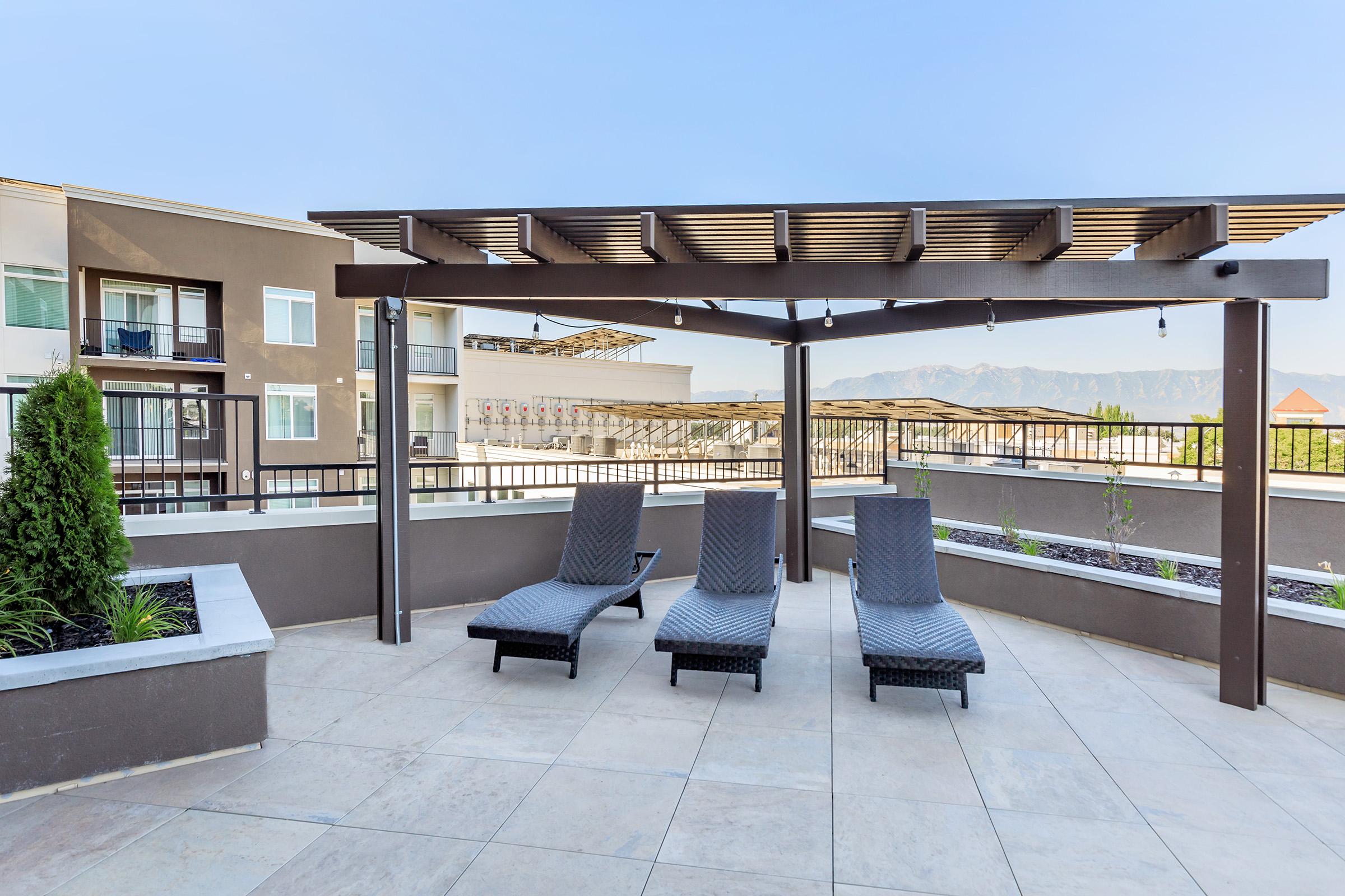 A modern rooftop patio featuring three lounge chairs under a stylish pergola. The space includes landscaped areas and overlooks a scenic view with mountains in the background, ideal for relaxation and enjoying outdoor living. Bright blue sky enhances the inviting atmosphere.