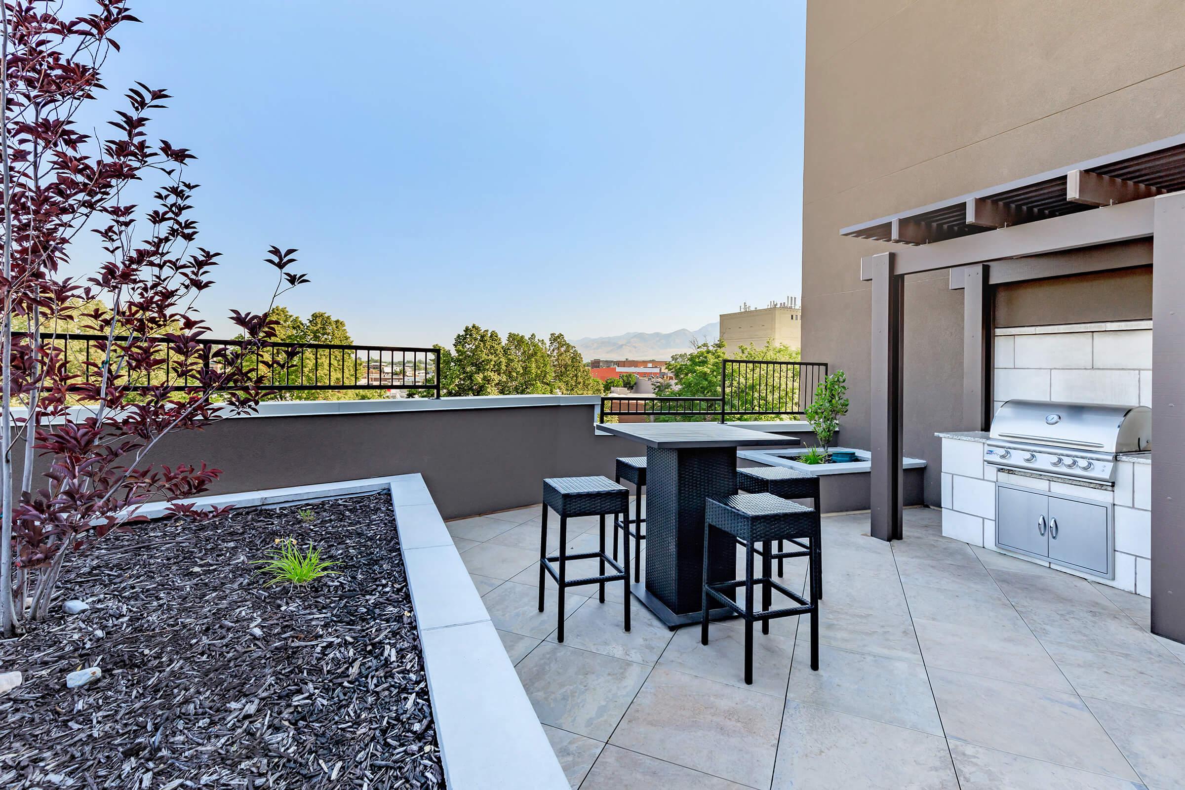 A spacious rooftop terrace featuring a modern outdoor kitchen with a grill, black bar stools, and a small table. Surrounding greenery adds a touch of nature, while a clear blue sky and distant mountains create a serene backdrop. The area is designed for relaxation and entertainment.