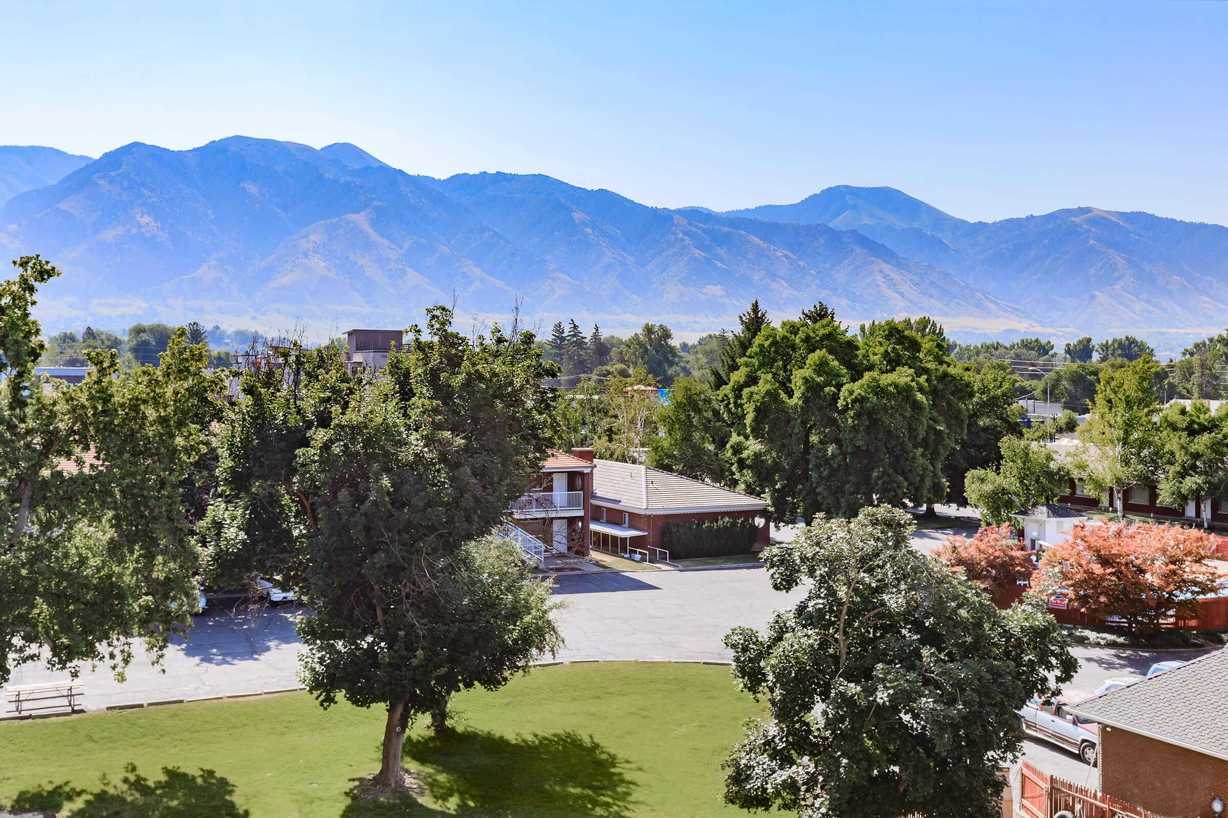 A scenic view of a mountain range under clear blue skies, with lush trees and a green lawn in the foreground. In the middle ground, there are several buildings, including residential-style architecture. The backdrop features rolling hills and distant mountains, showcasing a peaceful outdoor setting.