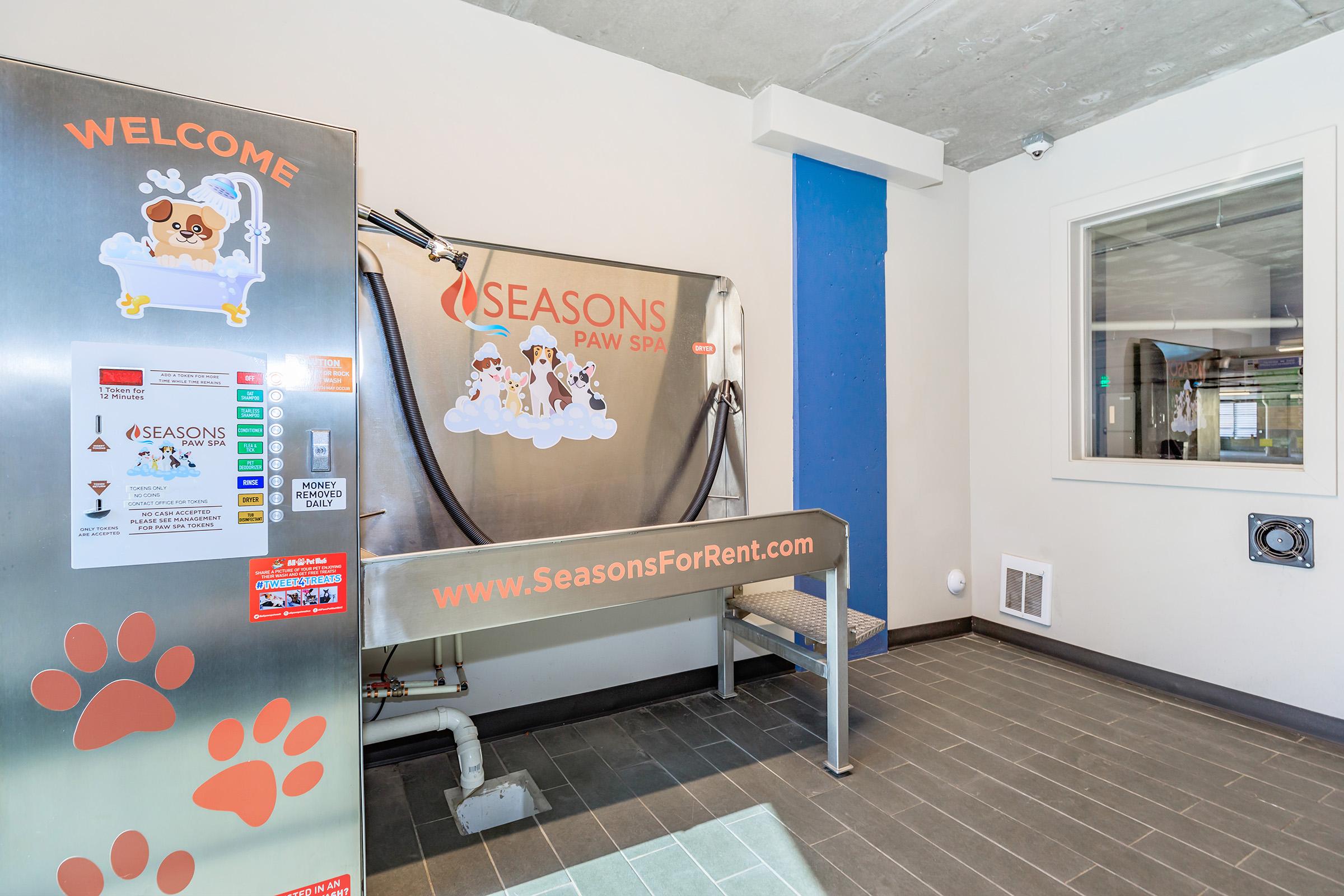 A pet grooming area featuring a stainless steel bathing station with a sign that reads "SEASONS PAW SPA." The wall is painted blue, and there are dog-themed decorations. A welcome sign is visible, along with various informational flyers or instructions on the grooming station. The space is well-lit and modern.