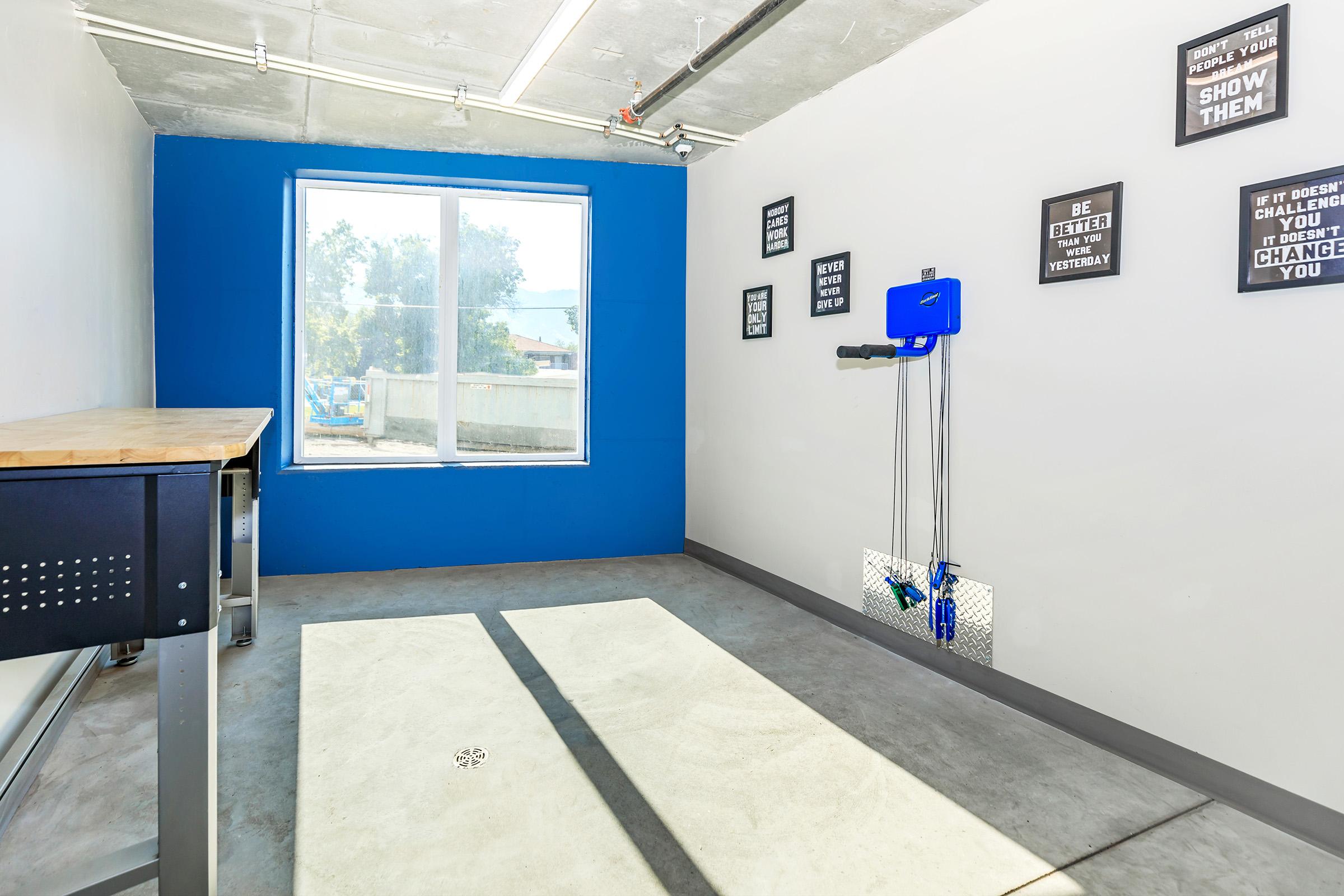 A bright, minimalist room with a blue accent wall, featuring a large window that lets in natural light. There are motivational posters on the walls, a wooden table in the corner, and exercise equipment attached to the wall, creating a functional space for workouts or activities. The floor is gray concrete.