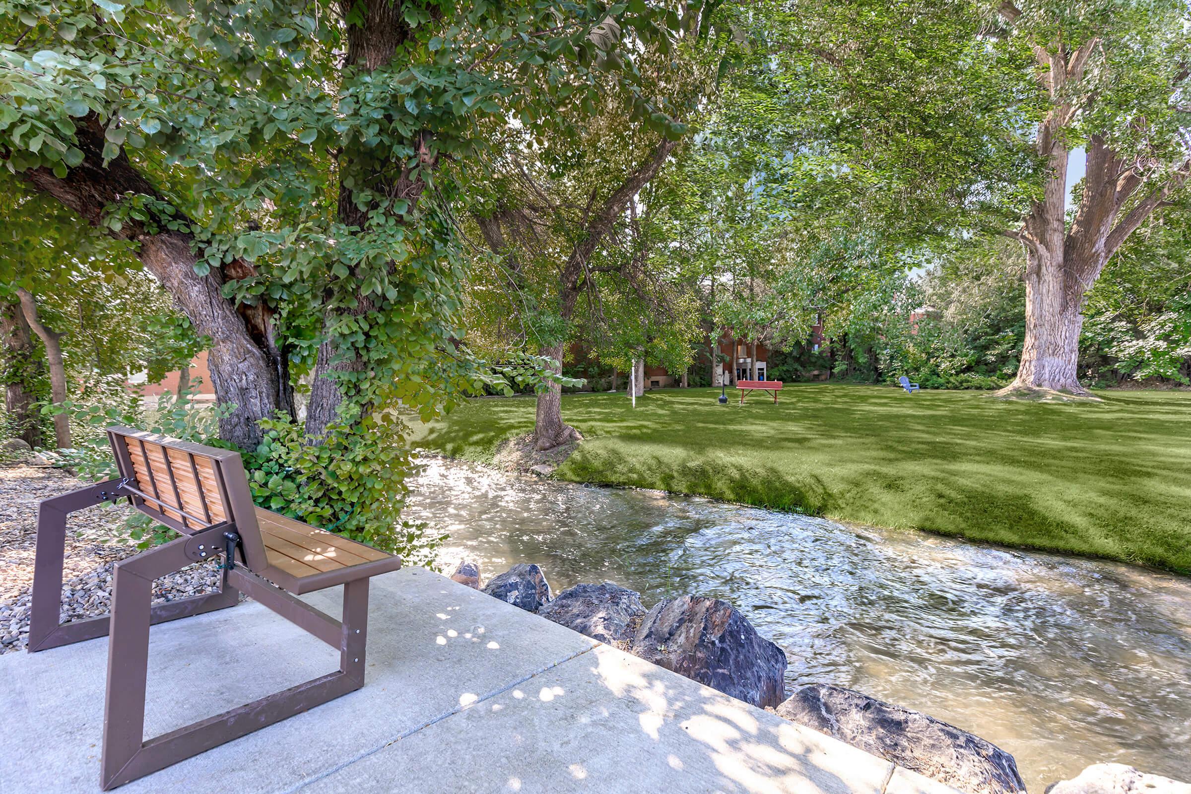 A peaceful park scene featuring a wooden bench beside a flowing stream. Lush green grass surrounds the area, with tall trees providing shade. In the background, a glimpse of a cozy seating area can be seen, enhancing the tranquil atmosphere of the outdoor space.