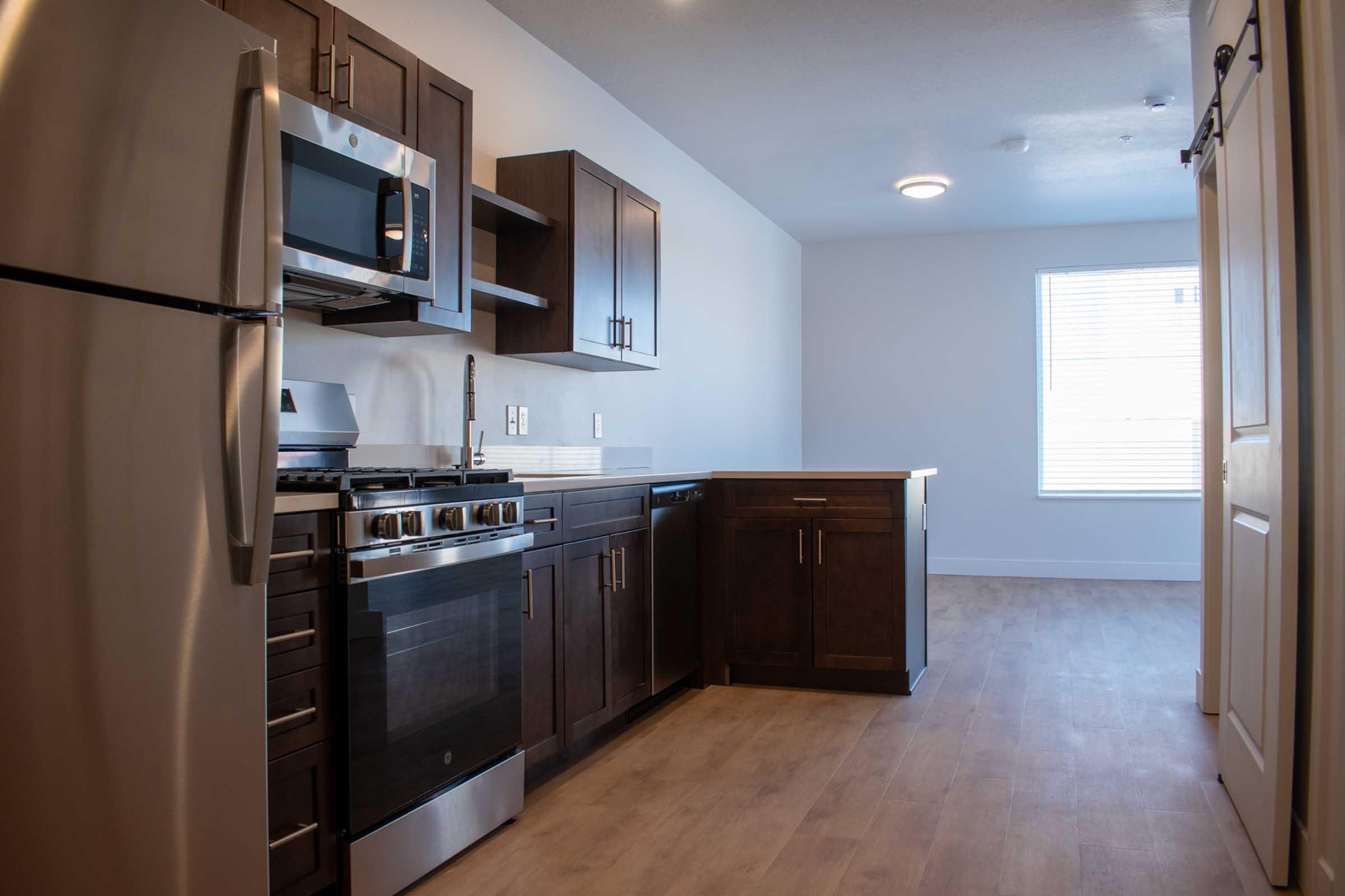 Modern kitchen featuring stainless steel appliances, including a microwave and oven, dark wooden cabinets, a countertop, and a window letting in natural light. The flooring is light wood, and the space appears clean and well-lit.