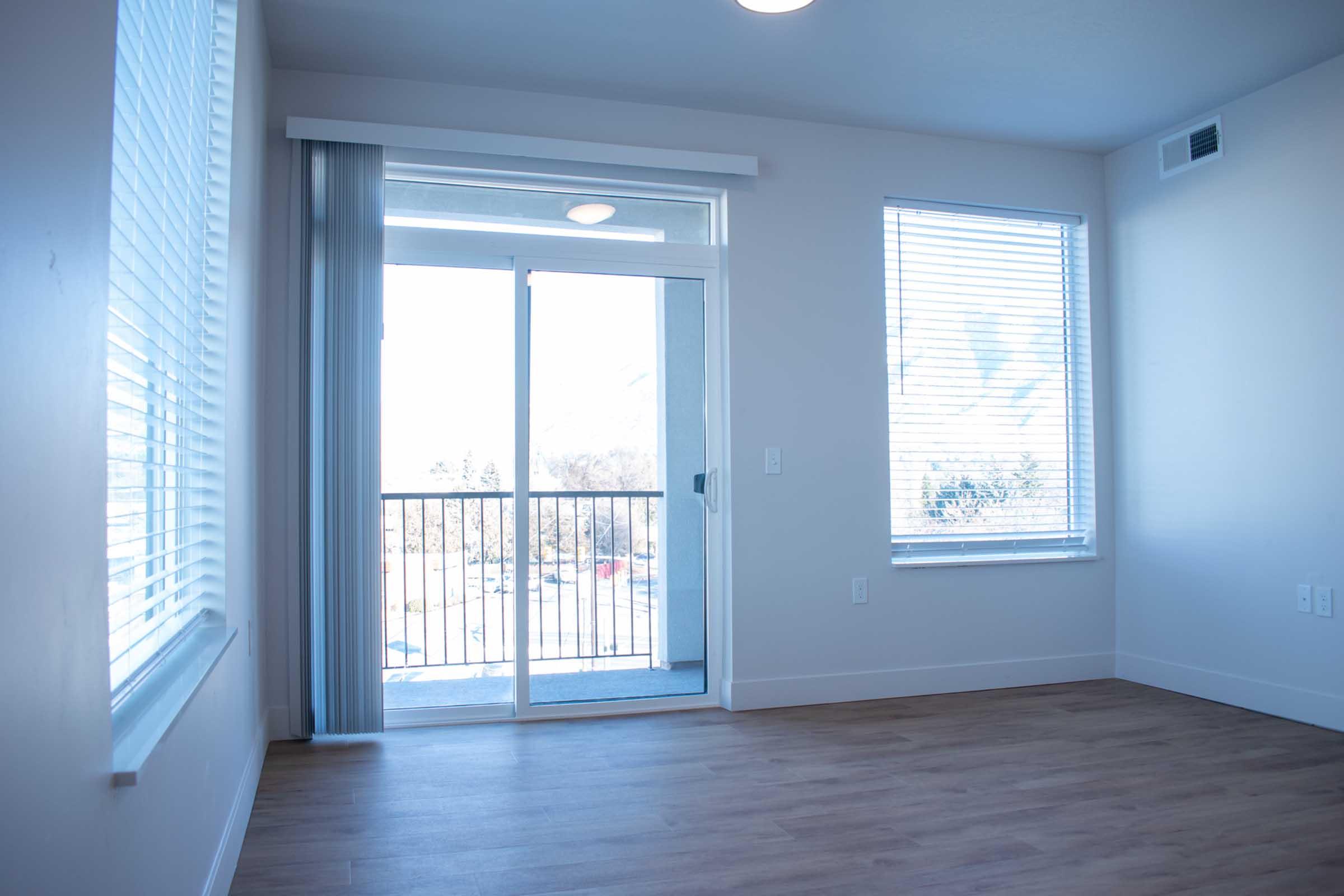 A bright, empty room featuring light-colored walls and natural wood flooring. There are large windows with blinds allowing sunlight to enter, and a sliding glass door that leads to a balcony. The room has a modern design, conveying a spacious and airy feel.