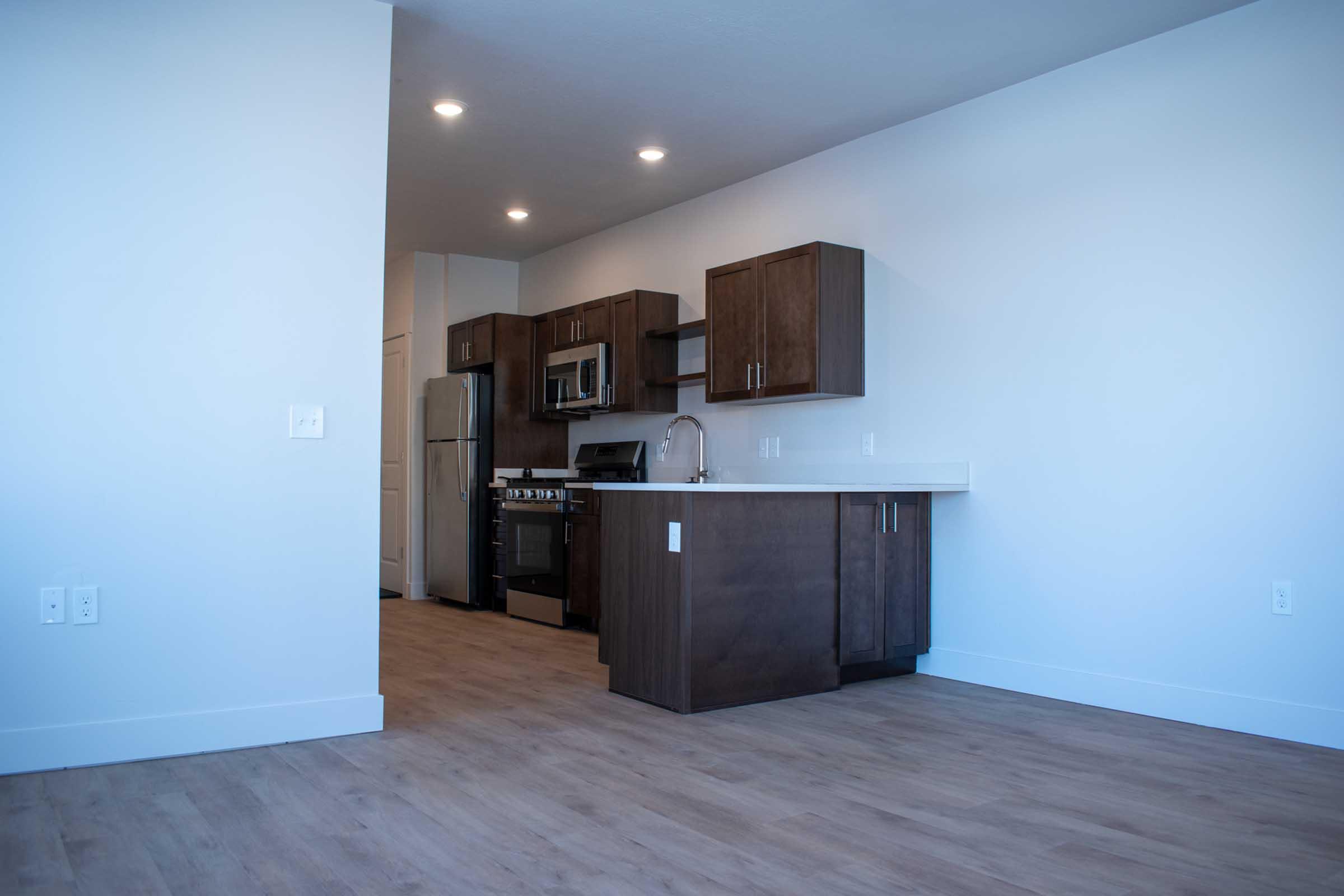 Modern kitchen with sleek dark wood cabinets and stainless steel appliances, featuring a silver refrigerator, microwave, and stove. The room has white walls and a light wood floor, creating a bright and spacious atmosphere. The kitchen island includes a sink and ample counter space.