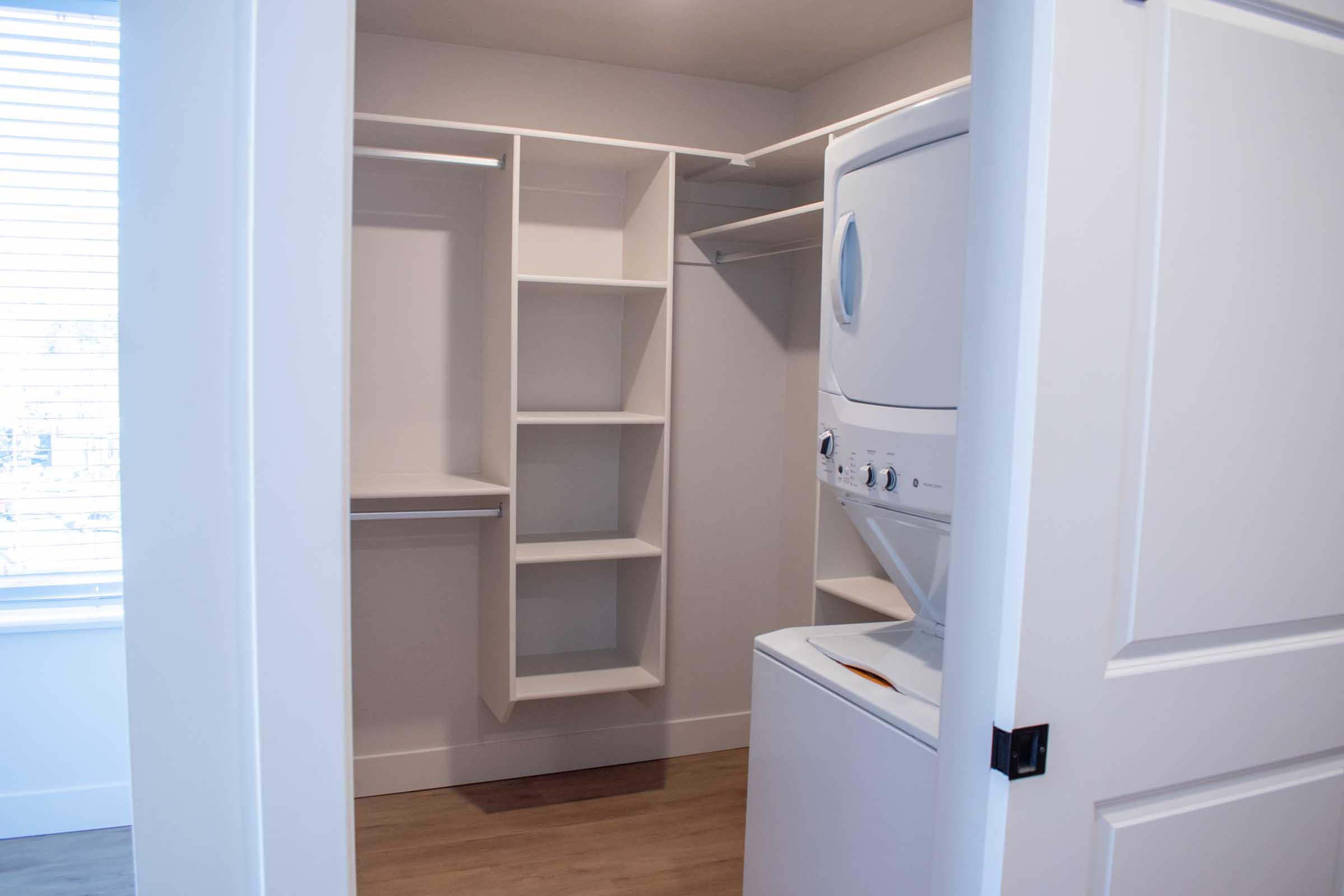 A modern laundry area featuring a stacked washer and dryer situated next to open shelving and rod for hanging clothes. The space has neutral walls, a door leading outside, and a window with blinds allowing natural light to enter. The flooring is light wood, creating a clean and bright atmosphere.