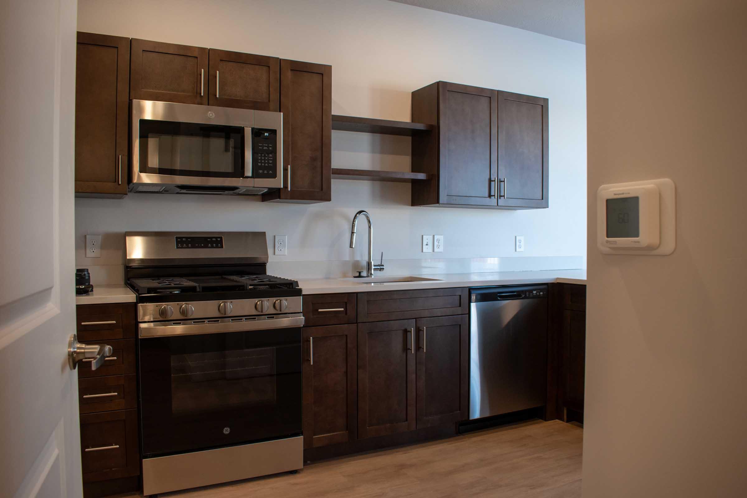 Modern kitchen with dark wooden cabinets and stainless steel appliances. Includes a gas stove, microwave, and dishwasher. Countertops are light-colored, creating a contrast with the cabinetry. A sink and open shelves are also visible. The room has a warm, inviting atmosphere with wooden flooring.