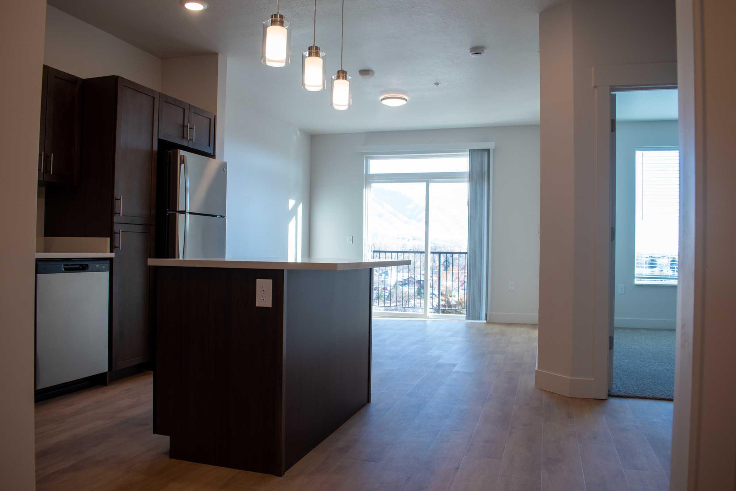 Modern kitchen and living area with an island countertop, stainless steel appliances, and large windows providing natural light and a view. The space features light-colored walls and hardwood flooring, with a balcony visible through the glass doors.