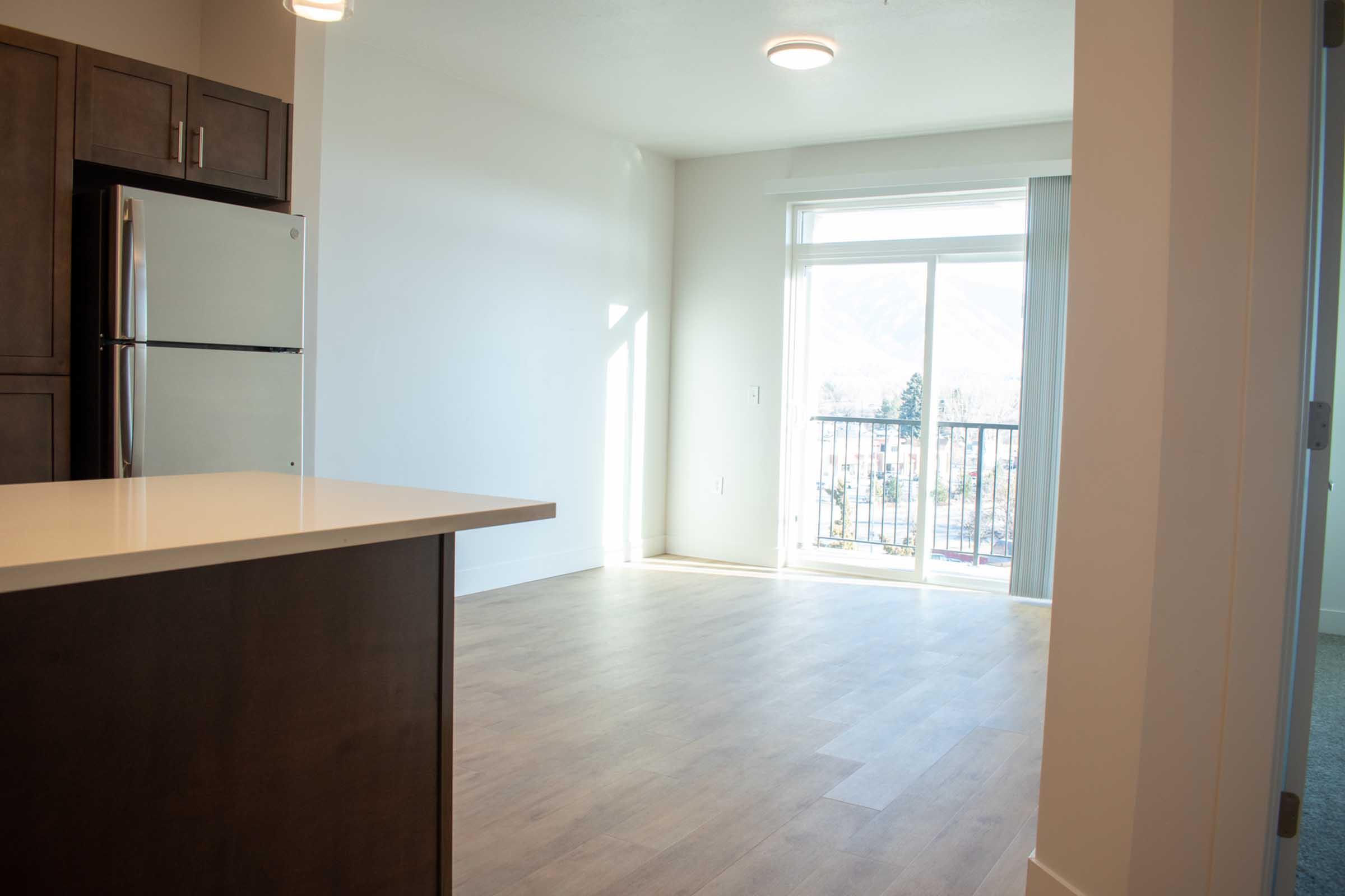 A bright, modern living space featuring a kitchen area with a stainless steel refrigerator and a countertop. The room has large windows with a view of a balcony and natural light streaming in. Neutral-colored walls and wood flooring create an open and inviting atmosphere.