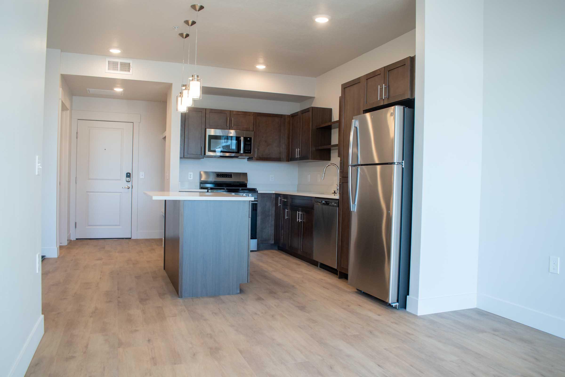 Modern kitchen in an apartment featuring stainless steel appliances, including a refrigerator and microwave, wooden cabinetry, and a large island. The floor is light-colored laminate, and the space is well-lit with recessed lighting. A door is visible leading to the main entrance.