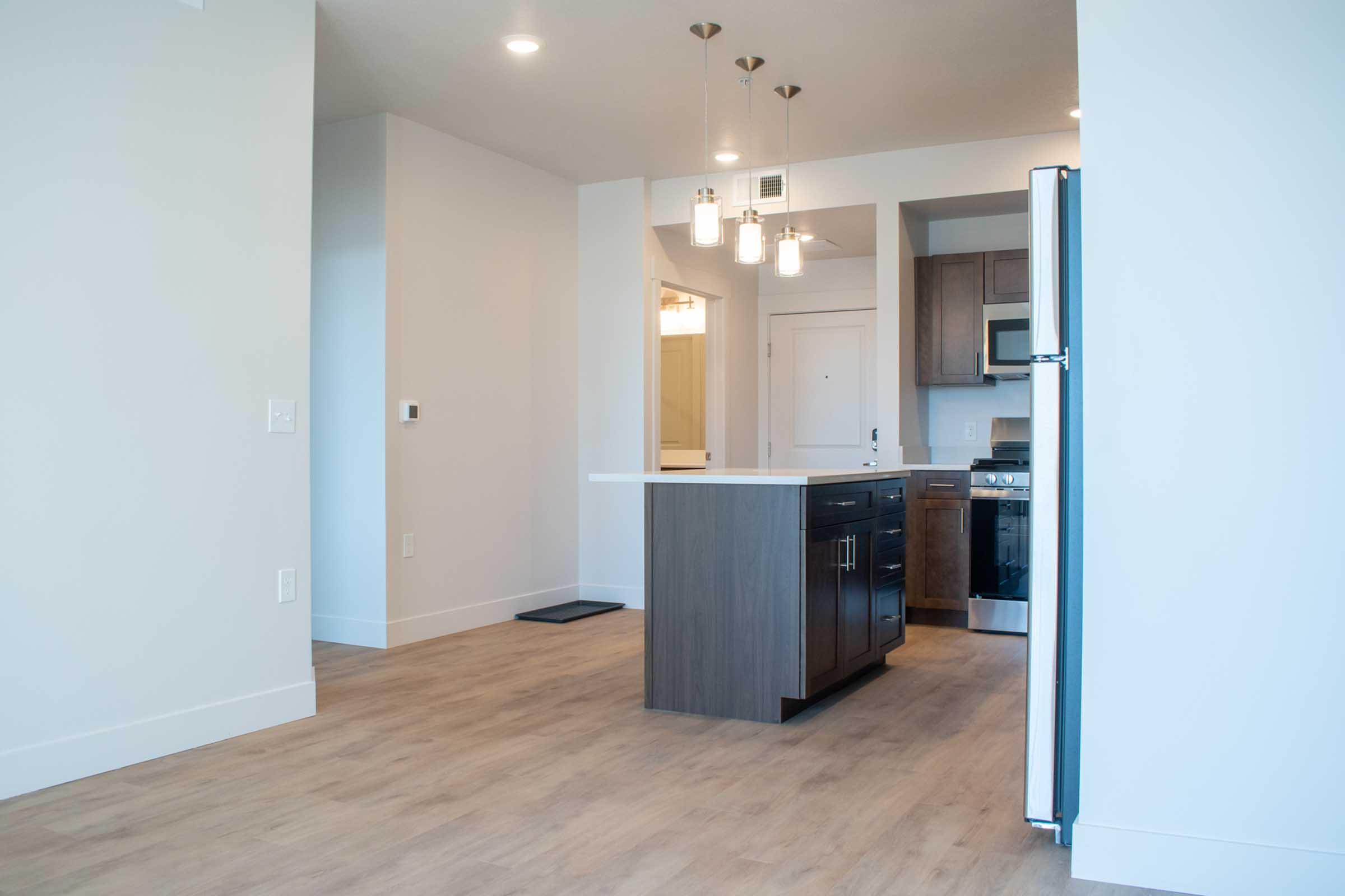 Modern kitchen interior featuring light wood flooring, an island with a dark cabinet, stainless-steel appliances, and pendant lighting. The space is bright and open, with minimalist design elements and a neutral color palette.