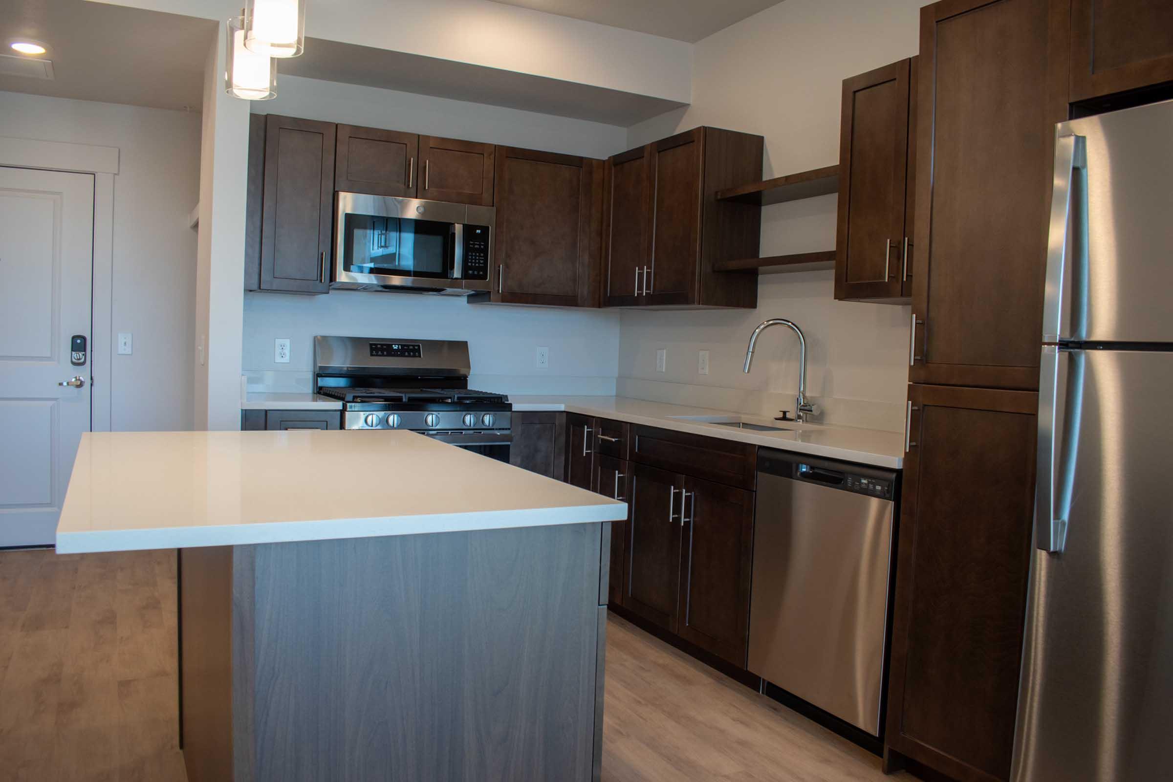 Modern kitchen featuring dark wooden cabinets, stainless steel appliances including a microwave, stove, and dishwasher. A white countertop island serves as a focal point. The space has neutral-colored walls and a bright, airy feel, enhanced by overhead light fixtures.