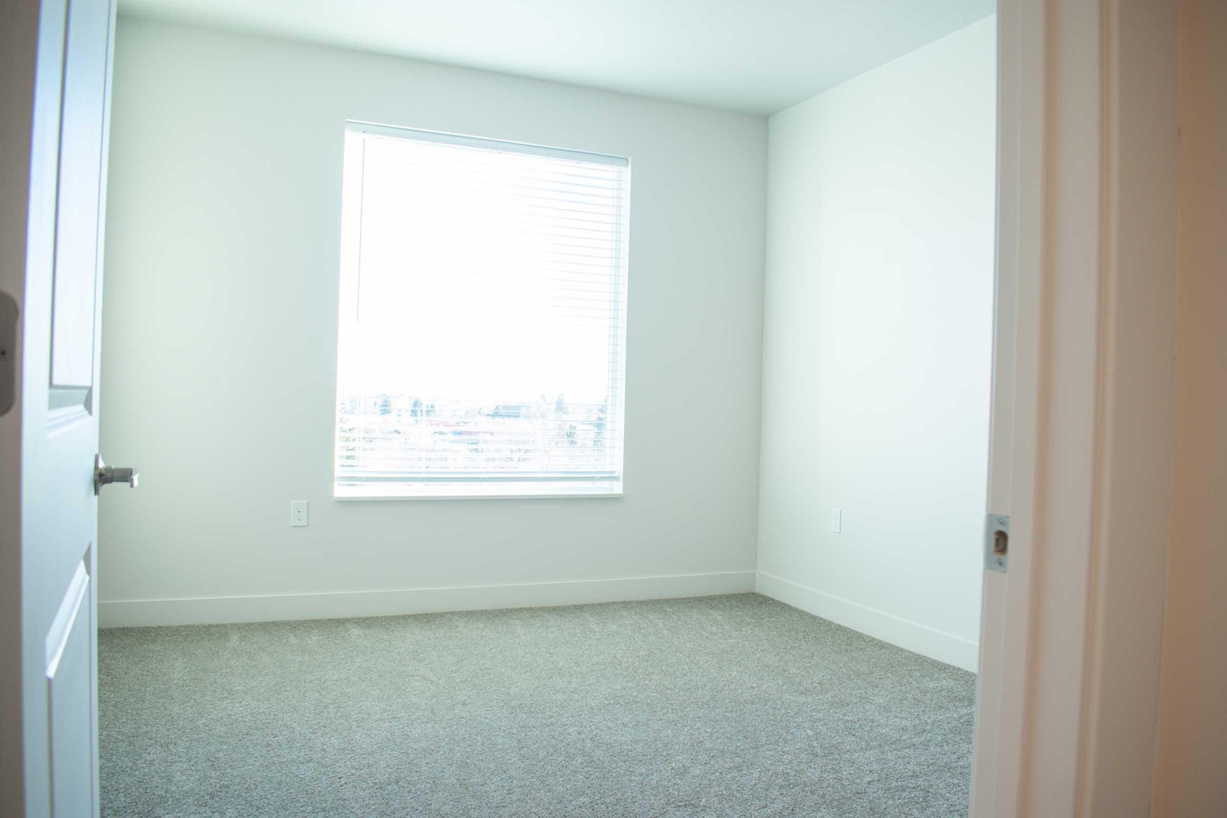 A bright, empty room featuring light-colored walls and a large window with blinds. The floor is covered in gray carpet, and the doorway is visible on the right side of the image, leading into the room. Natural light filters in through the window, illuminating the space.