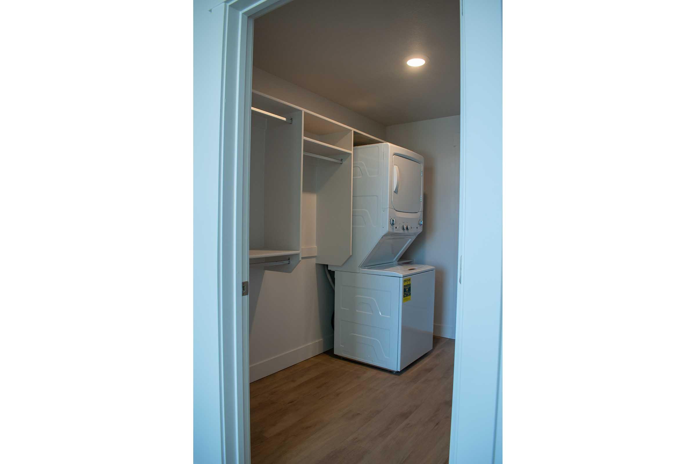 A laundry room with a stacked washer and dryer, white cabinetry, and light-colored wood flooring. The room is well-lit with a ceiling light, and there is an open space for hanging clothes on the left. The walls are painted in a neutral color, providing a modern and clean aesthetic.