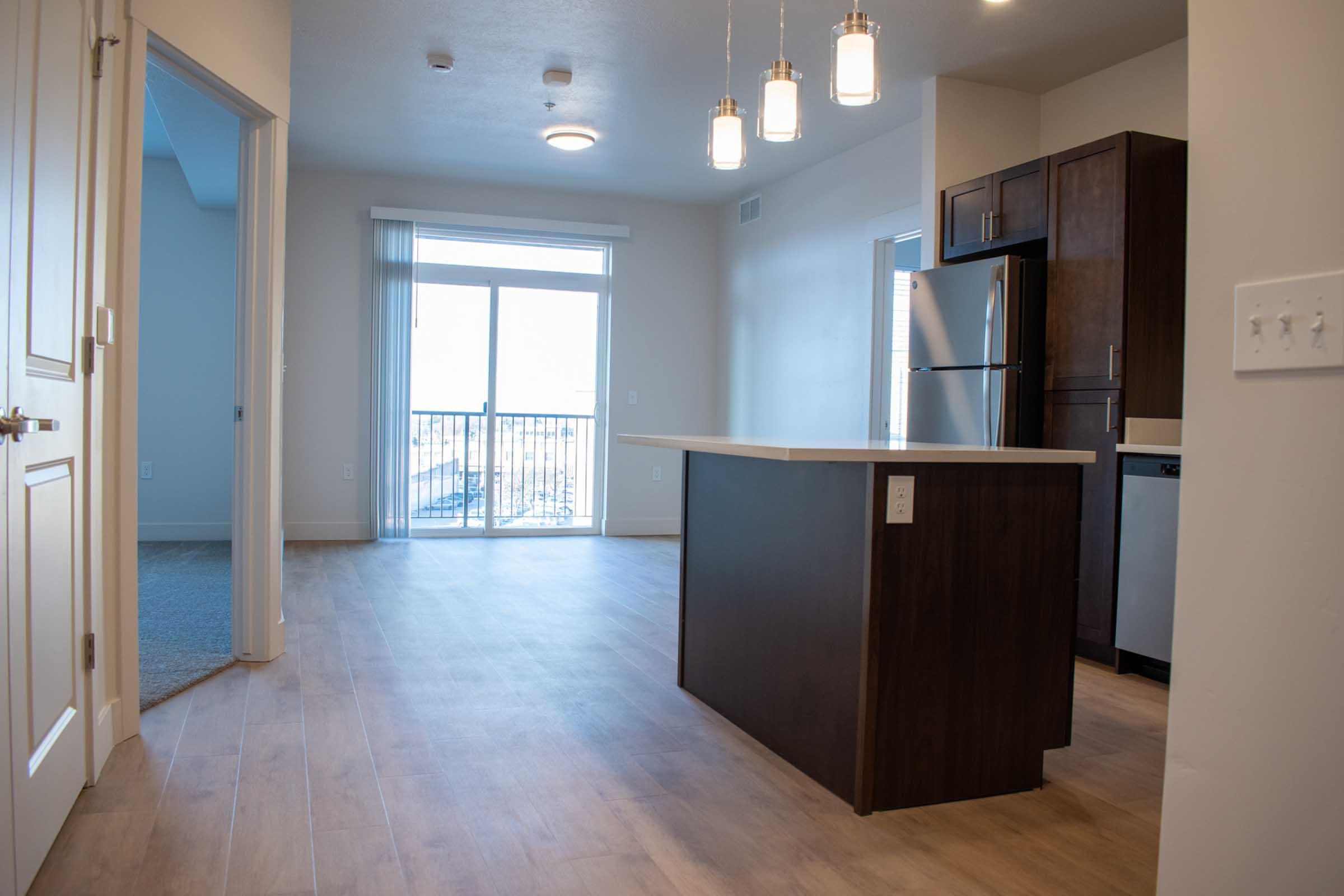 Spacious modern kitchen with a central island, featuring pendant lights. Open layout leads to a living area with large windows and a balcony door, allowing natural light. Neutral-colored walls and wooden flooring create a warm atmosphere. Stainless steel appliances are partially visible in the kitchen.