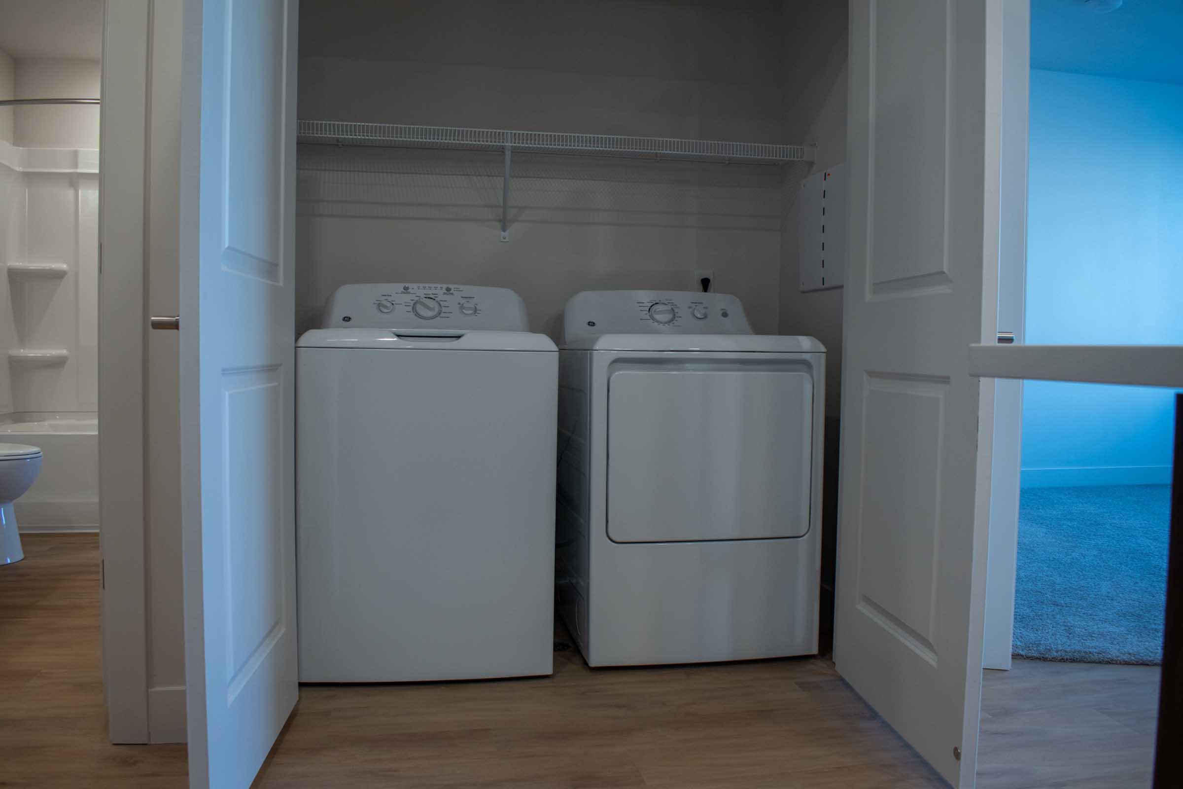 A laundry area featuring a white washing machine and dryer side by side, positioned in a bright, tidy space. The room has light-colored walls and a closet-like appearance with doors partially open. A glimpse of a bathroom can be seen in the background.