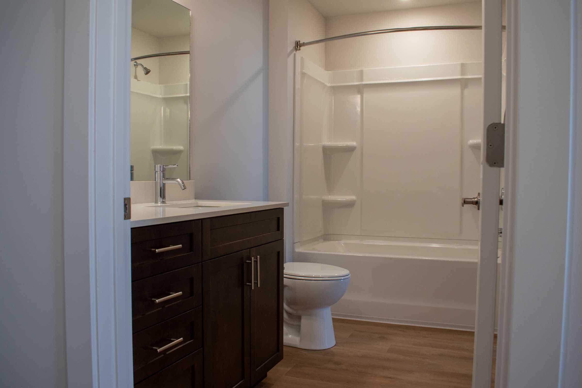 A modern bathroom featuring a shower-tub combo, a dark wood vanity with multiple drawers, a rectangular sink, a large mirror, and wooden flooring. The space is well-lit and has a clean, minimalist design.