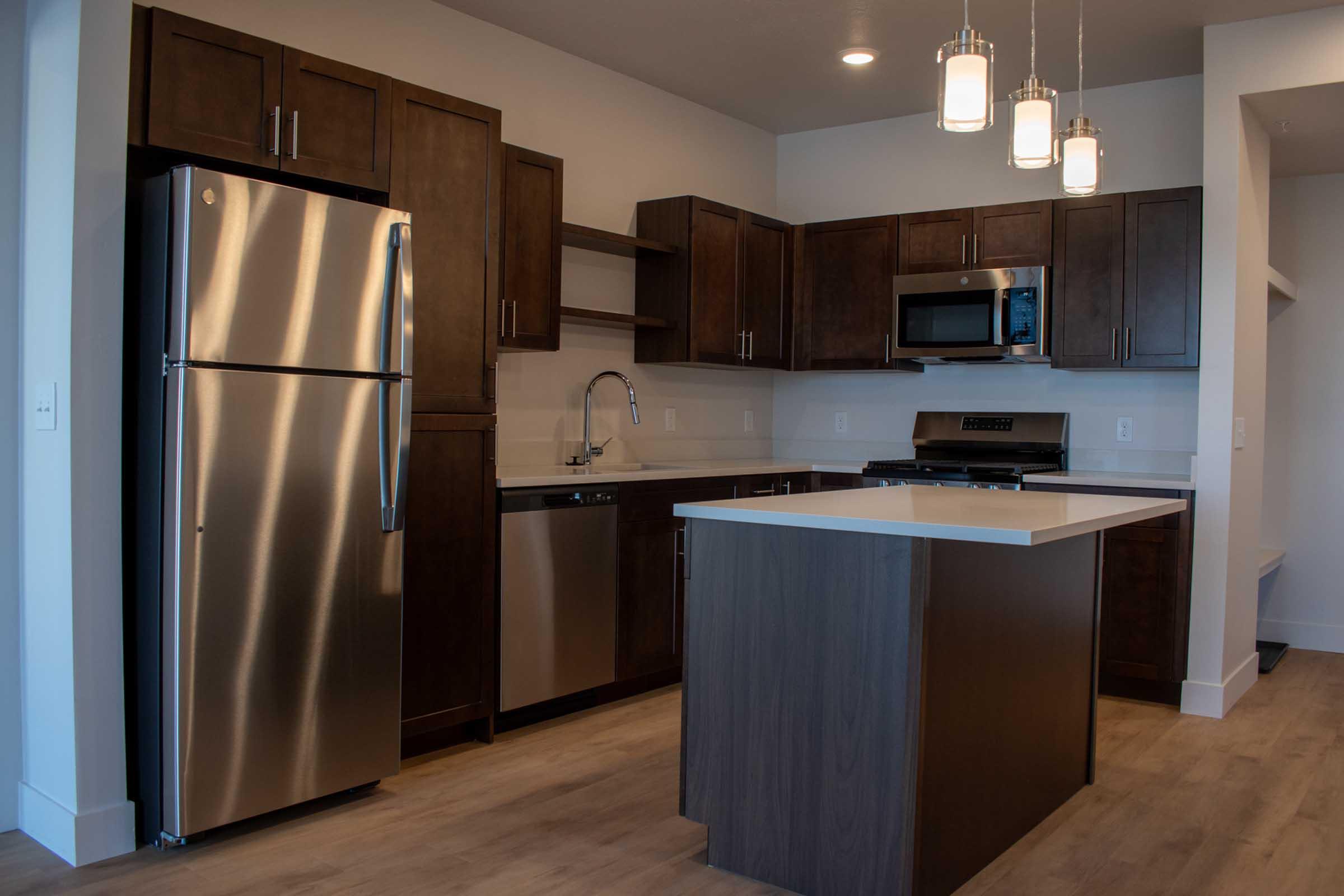 A modern kitchen featuring dark wood cabinetry, stainless steel appliances including a refrigerator, dishwasher, and microwave, a gas stove, and a sleek island with a white countertop. The room is well-lit with pendant lights and has a contemporary design.