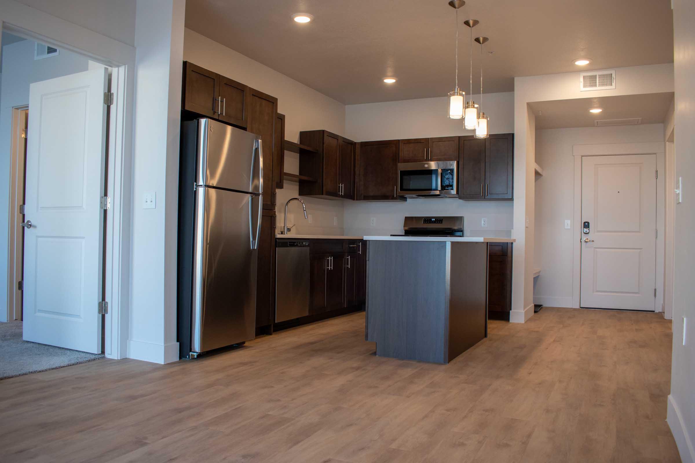 Modern kitchen featuring dark wood cabinetry, stainless steel appliances, and an island with pendant lighting. The space includes a refrigerator, microwave, and dishwasher, with light-colored walls and a wood-like floor. A doorway leads to an entrance hall.