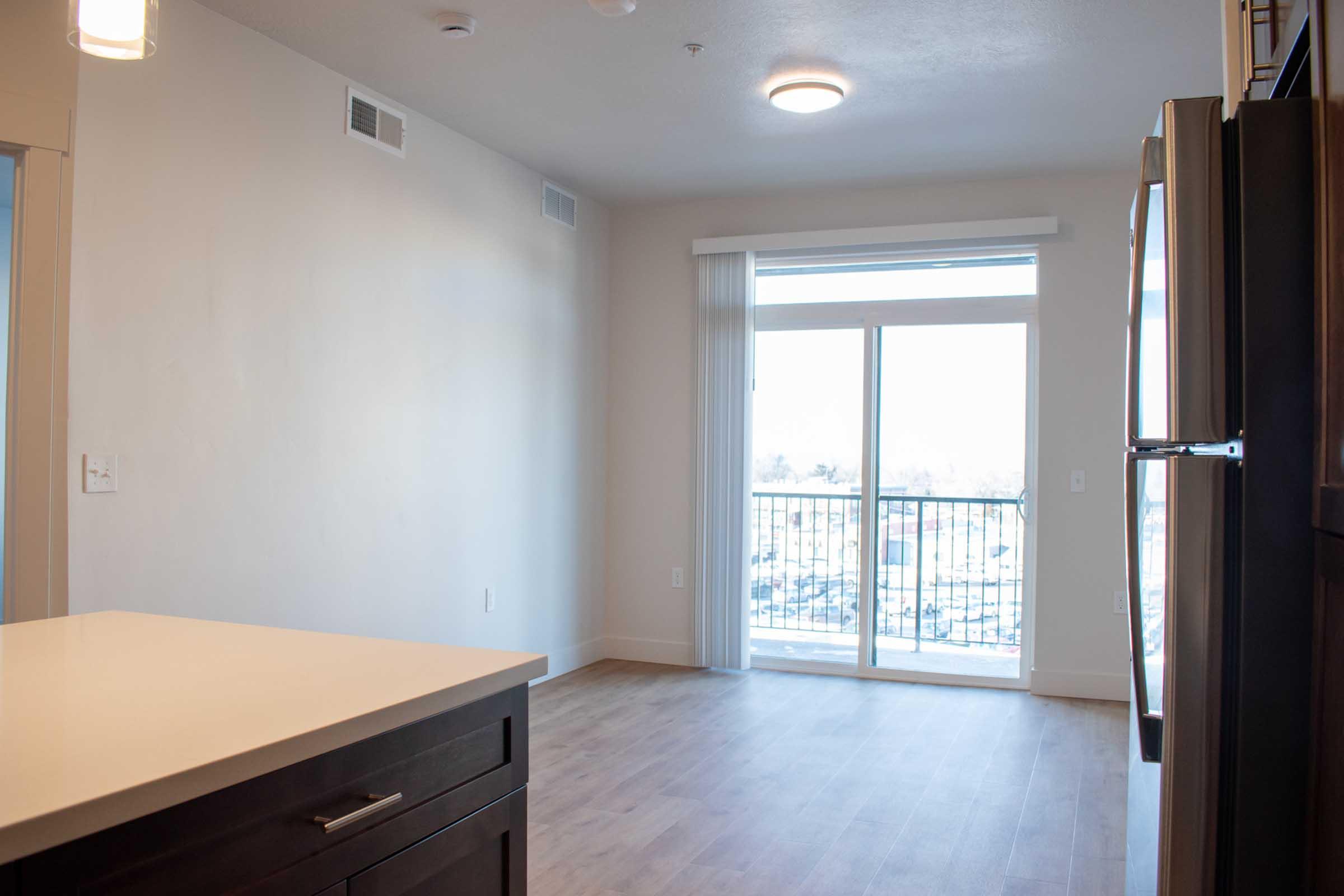 A bright, modern living space featuring a light-colored wall and large sliding glass door that leads to a balcony. The room includes a kitchen island with dark cabinetry, wooden flooring, and a ceiling light fixture. Natural light streams in from the balcony, creating an inviting atmosphere.