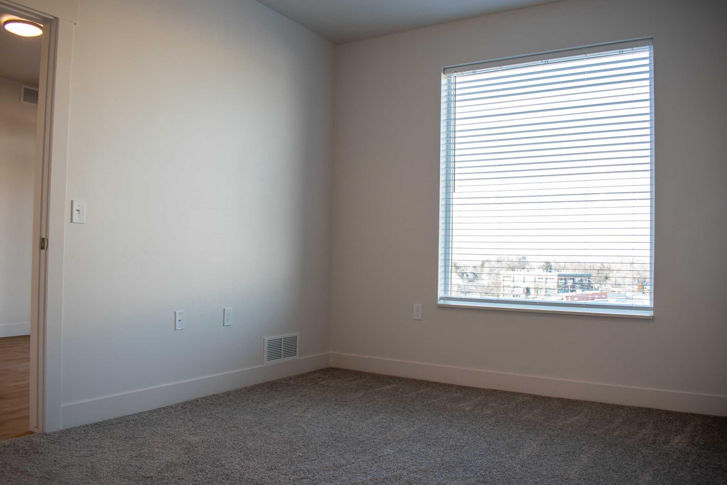 A bright, empty room with light-colored walls and a window covered by white blinds. The floor is carpeted in a light gray color. There is a door on the left side of the image and a vent on the wall below the window. The room appears clean and spacious, with natural light coming through the window.