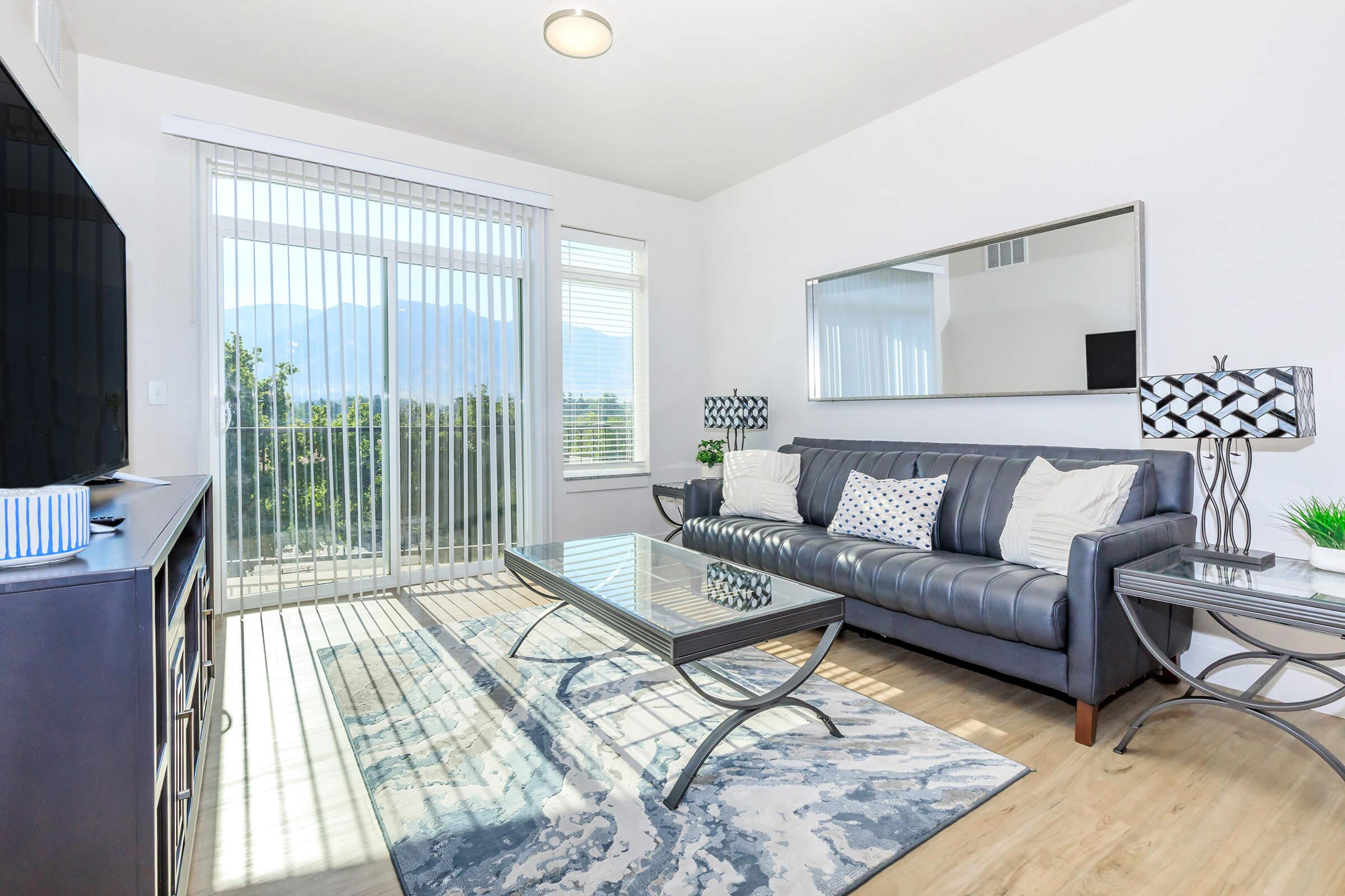 Modern living room featuring a gray sofa with decorative pillows, a glass coffee table, and two stylish table lamps. Large sliding glass doors allow natural light and showcase a mountain view. The space has light wood flooring and a decorative rug, enhancing the contemporary aesthetic.